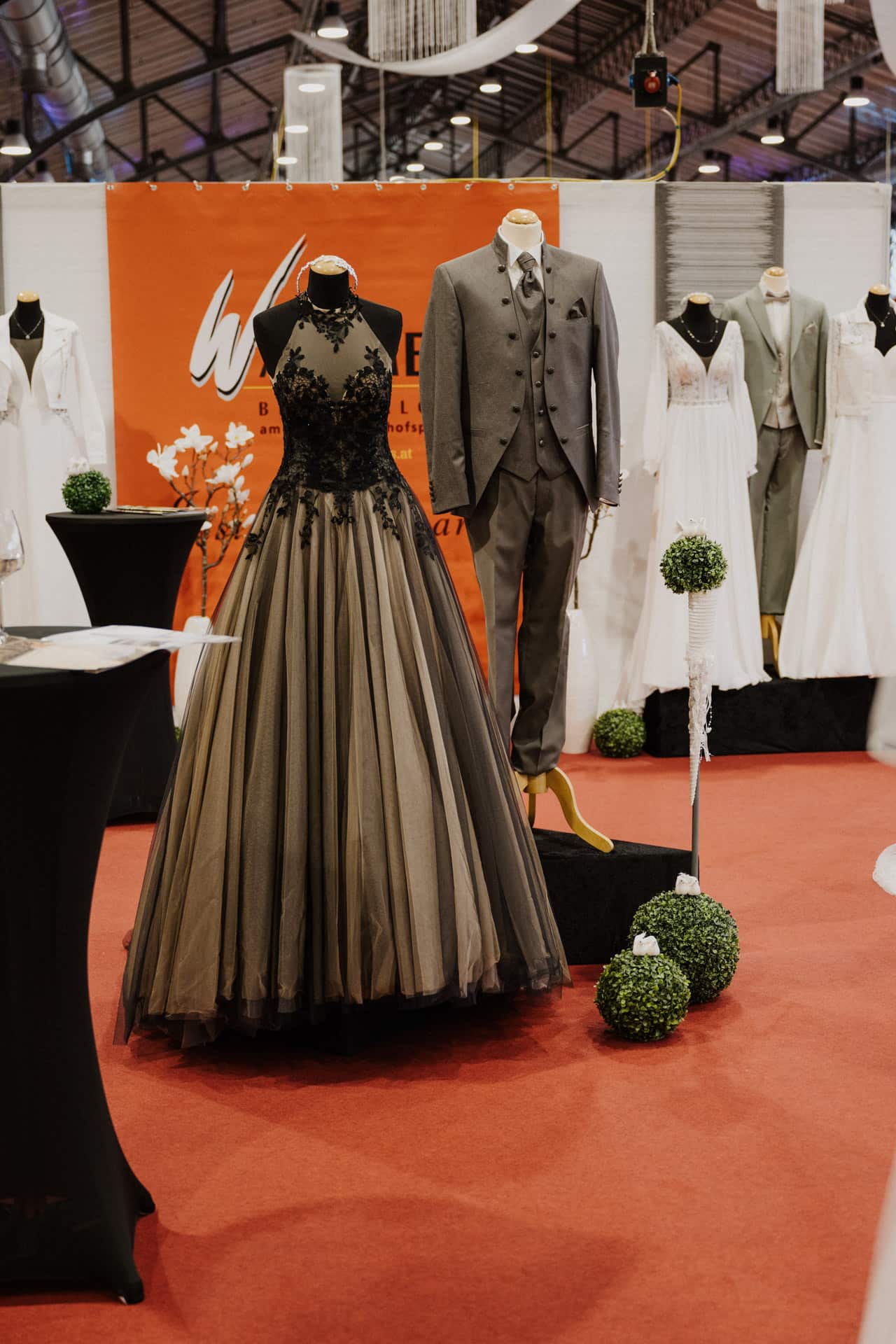 Eine Ausstellung von Abendgarderobe auf Schaufensterpuppen, darunter ein schwarz-graues Abendkleid aus Spitze mit weitem Rock und ein grauer Anzug mit Weste und Krawatte. Umgeben sind weitere weiße Kleider vor einem Hintergrund mit Hochzeitsdekor, perfekt für Hochzeiten und Feste.