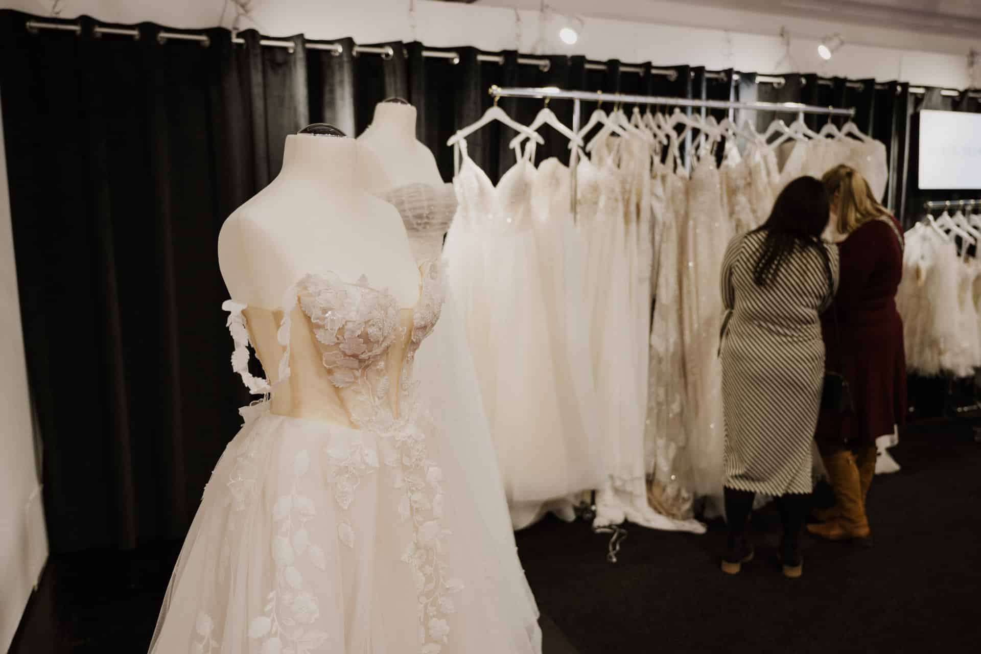 Zwei Schaufensterpuppen präsentieren elegante, weiße Brautkleider mit Blumenmuster, perfekt für Hochzeiten. Im Hintergrund hängen weitere Kleider an einer Kleiderstange. Zwei Frauen begutachten die Kleider und stehen neben der Kleiderstange in einer Boutique-Umgebung voller festlichem Charme.