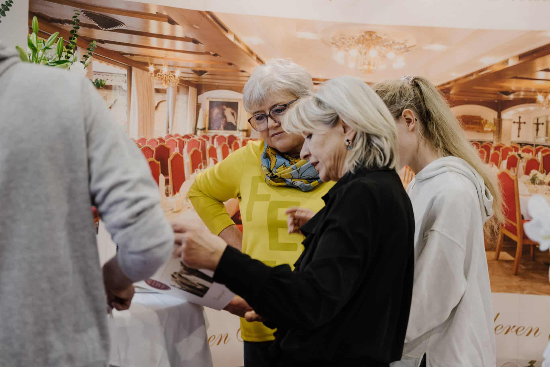 Drei Frauen stehen bei einer Indoor-Veranstaltung zusammen und prüfen ein Dokument. Die Kulisse, perfekt für Hochzeiten & Feste, ist mit einem großen Saal mit roten Stühlen dekoriert. Sie scheinen in ein Gespräch vertieft zu sein.