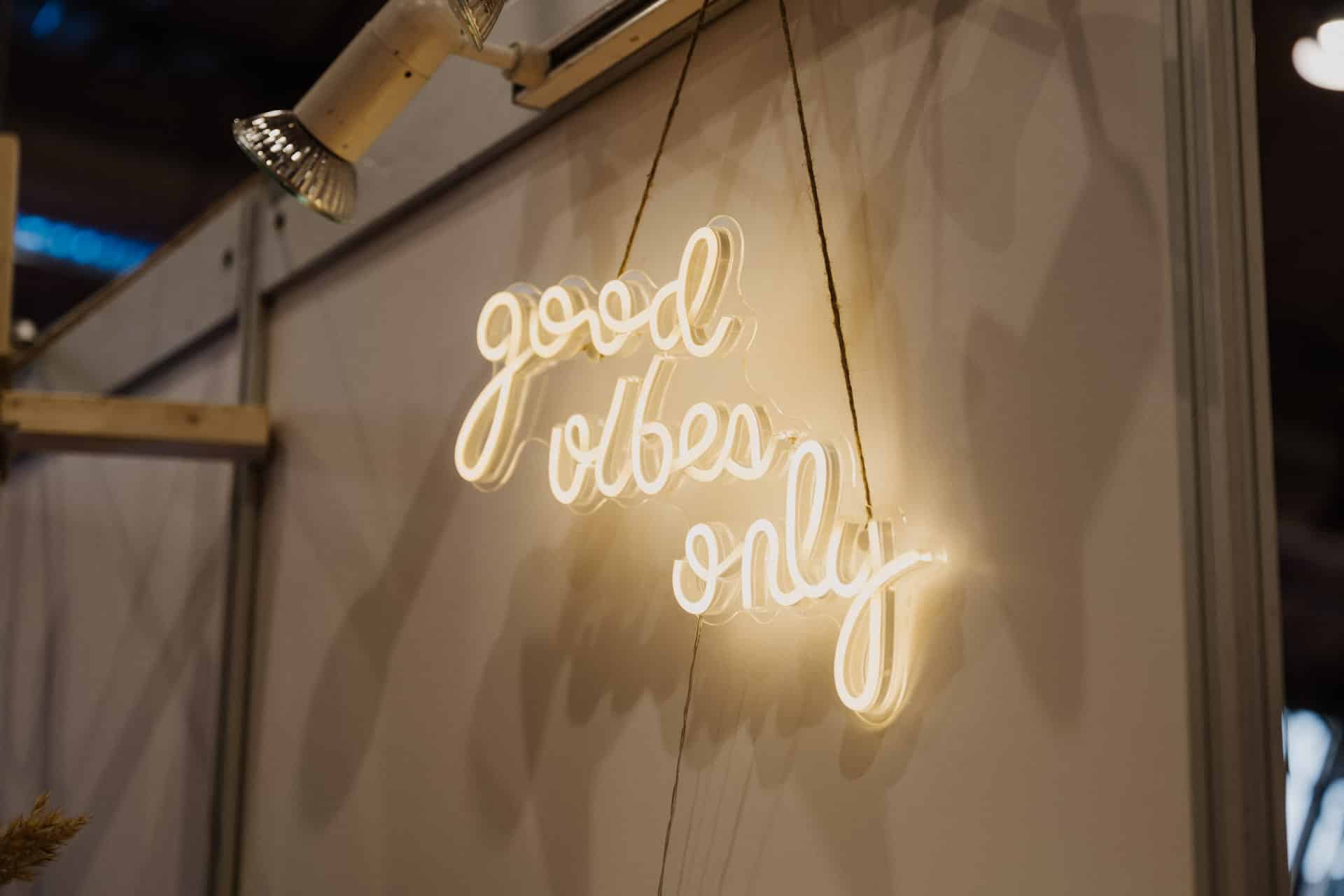 Auf einem warm beleuchteten Neonschild an einer Wand steht in Schreibschrift „good vibes only“ und verbreitet ein bezauberndes Licht, das perfekt für Hochzeiten ist. Der umgebende Bereich ähnelt einem eleganten Stand oder einem Ausstellungsaufbau mit einem Scheinwerfer darüber und schafft eine einladende Atmosphäre für festliche Zusammenkünfte.