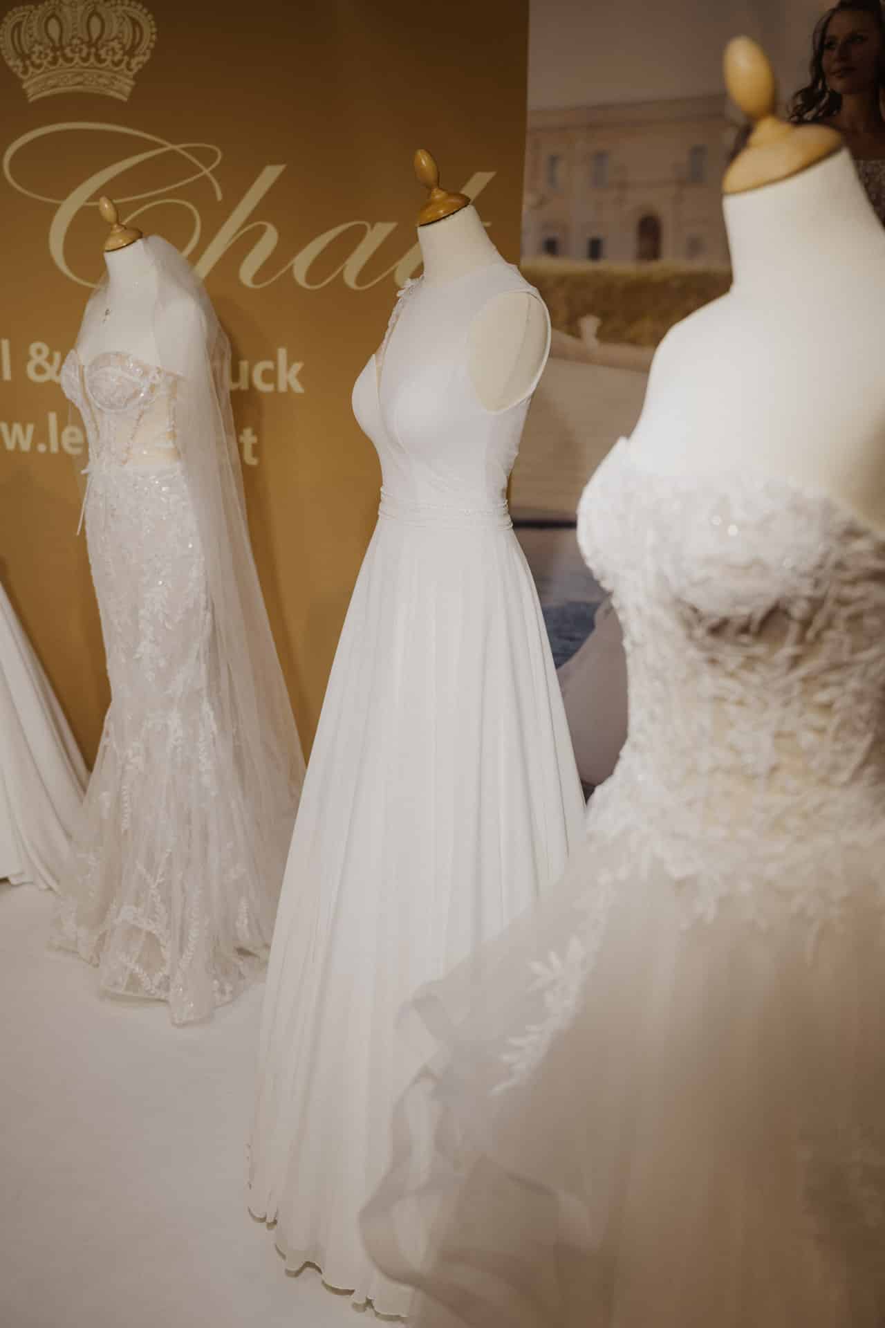 Eine Ausstellung eleganter weißer Brautkleider auf Puppen für Hochzeiten. Die Kleider sind in verschiedenen Stilen erhältlich, darunter Spitzendetails und weite Röcke. Hinter den Kleidern ist ein goldener Hintergrund mit teilweisem Text zu sehen, der für eine festliche Note sorgt.