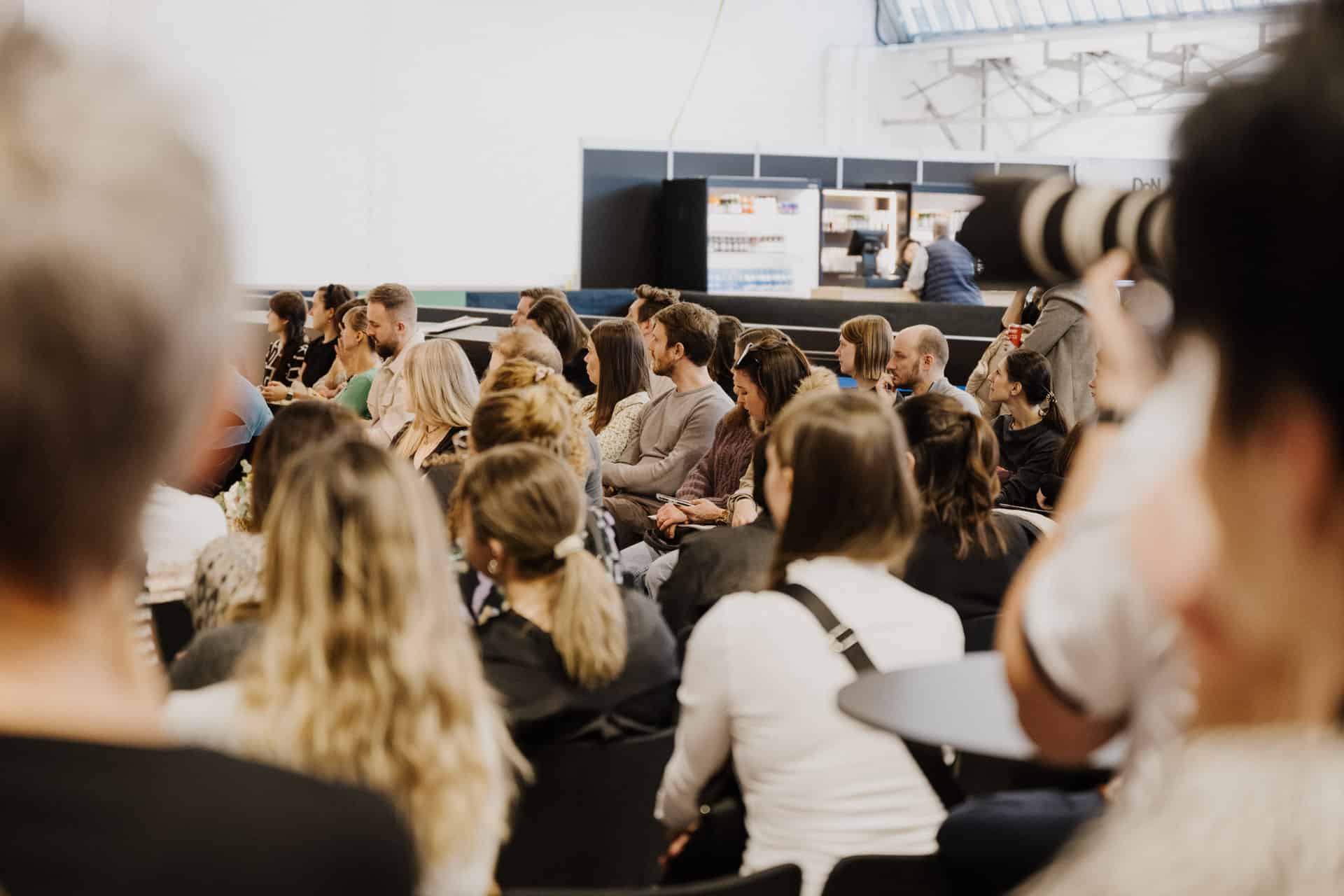 Das Publikum sitzt in einem Innenraum und hört aufmerksam zu. Es sind viele verschiedene Menschen anwesend, einige machen sich Notizen, andere sind in die Worte des Redners vertieft, was an Zusammenkünfte bei Hochzeiten & Festen erinnert. Im Vordergrund ist ein großes Kameraobjektiv zu sehen, das jeden Moment festhält.