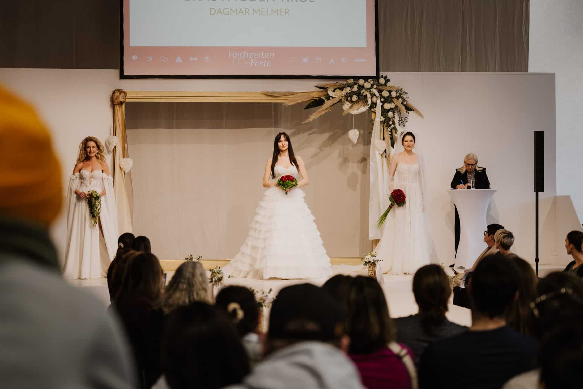 Drei Models in weißen Brautkleidern stehen auf einer Bühne, jedes hält einen Blumenstrauß in der Hand. Das Publikum schaut gespannt zu und ist fasziniert von der Eleganz, die perfekt für Hochzeiten ist. Rechts steht ein Mann an einem Podium. Im Hintergrund ist „DAGMAR RELMER“ zu sehen, und Blumenschmuck verstärkt die festliche Atmosphäre.