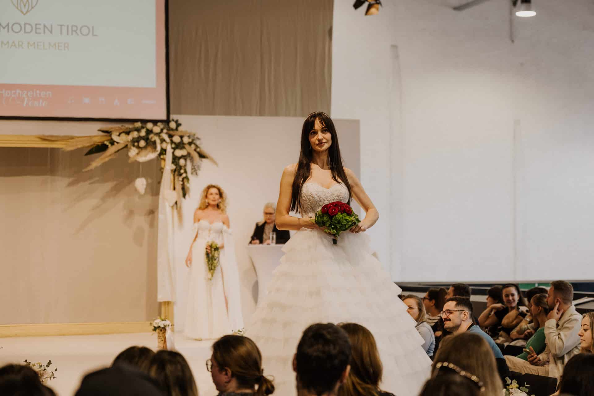 Eine Braut in einem gestuften weißen Brautkleid, einen Strauß roter und weißer Blumen in der Hand, schreitet anmutig über den Laufsteg bei diesem Hochzeitsspektakel. Eine weitere Braut in einem ähnlichen Kleid folgt und verzaubert das Publikum, das sich zu dieser festlichen Feier in einem wunderschön beleuchteten Veranstaltungsort versammelt hat.