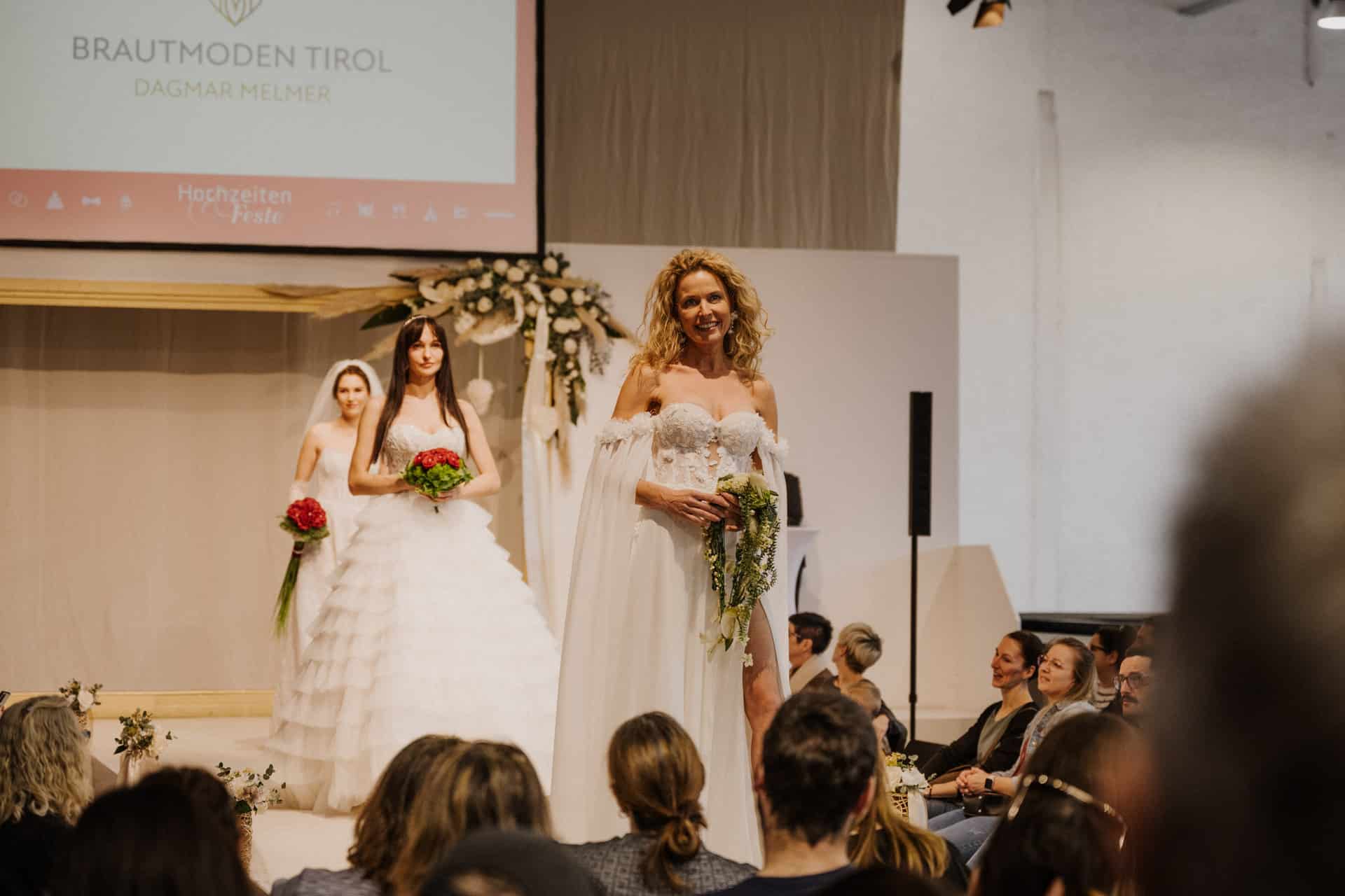 Eine Frau in einem weißen schulterfreien Kleid schreitet anmutig über den Laufsteg von Hochzeiten und hält dabei einen Blumenstrauß in der Hand. Zwei weitere Models in Brautkleidern stehen hinter ihr. Das Publikum sitzt auf beiden Seiten und ist von der festlichen Show fasziniert.