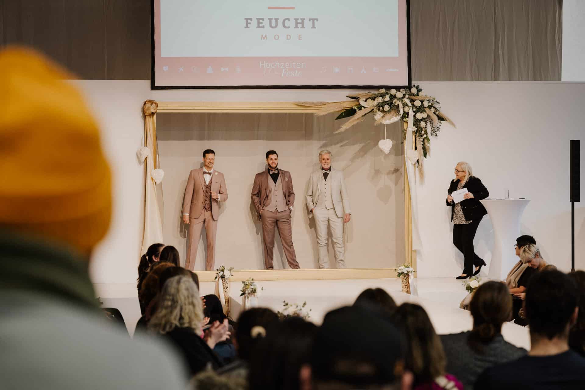 Drei Models in Anzügen stehen auf der Bühne unter einem mit Blumen geschmückten Holzbogen bei einer Modenschau, perfekt für Hochzeiten & Feste. Rechts steht ein Redner, im Vordergrund sitzt das Publikum. Oben ist auf einem Bildschirm „FEUCHT MODE“ zu sehen.