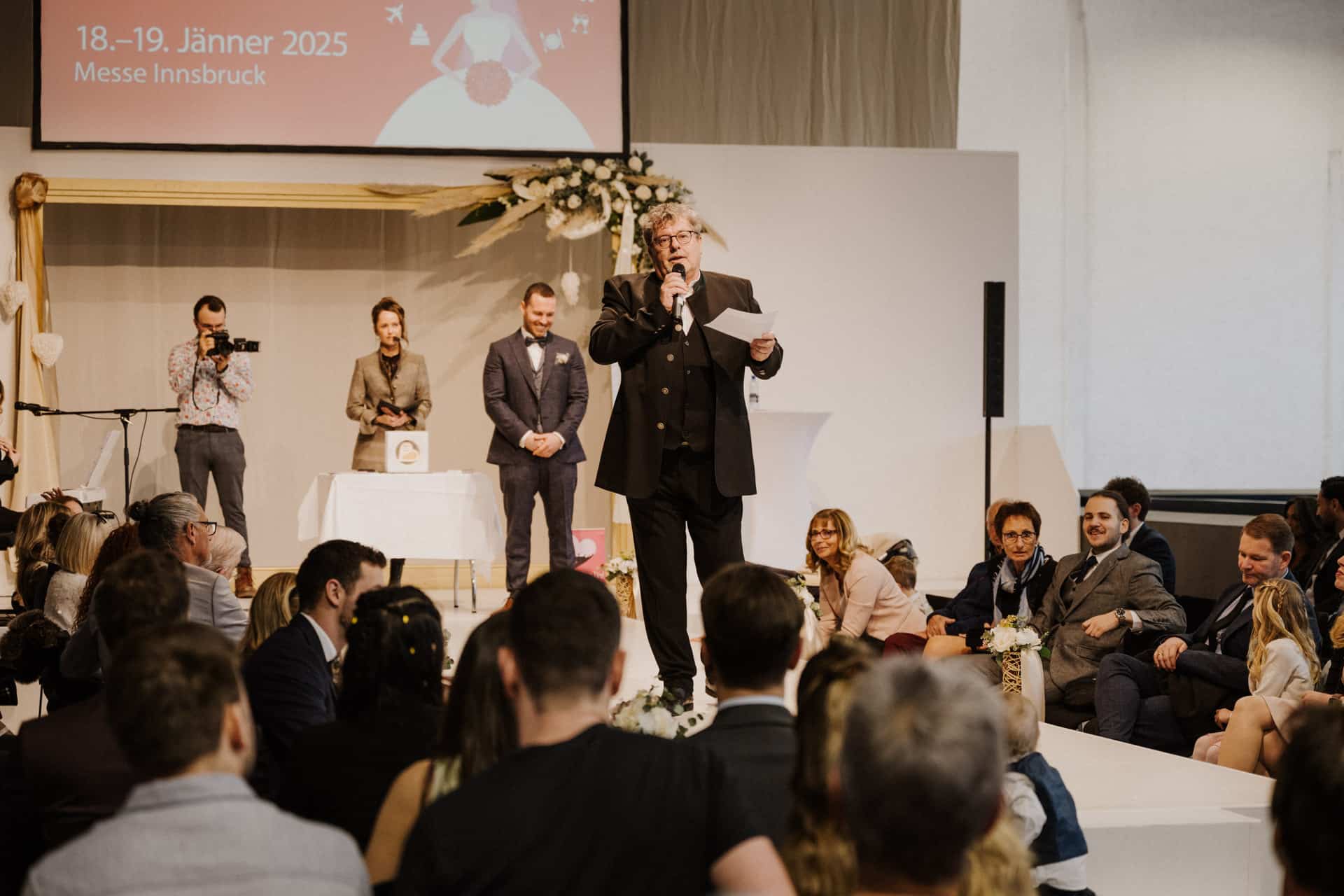 Bei einer Veranstaltung zu „Hochzeiten & Feste“ spricht ein Redner zu einem sitzenden Publikum. Ein Fotograf hält den Moment fest. Die Bühne ist mit Blumen geschmückt, auf einer Leinwand steht „18.-19. Jänner 2025, Messe Innsbruck“. Die Anwesenden sind engagiert und aufmerksam.