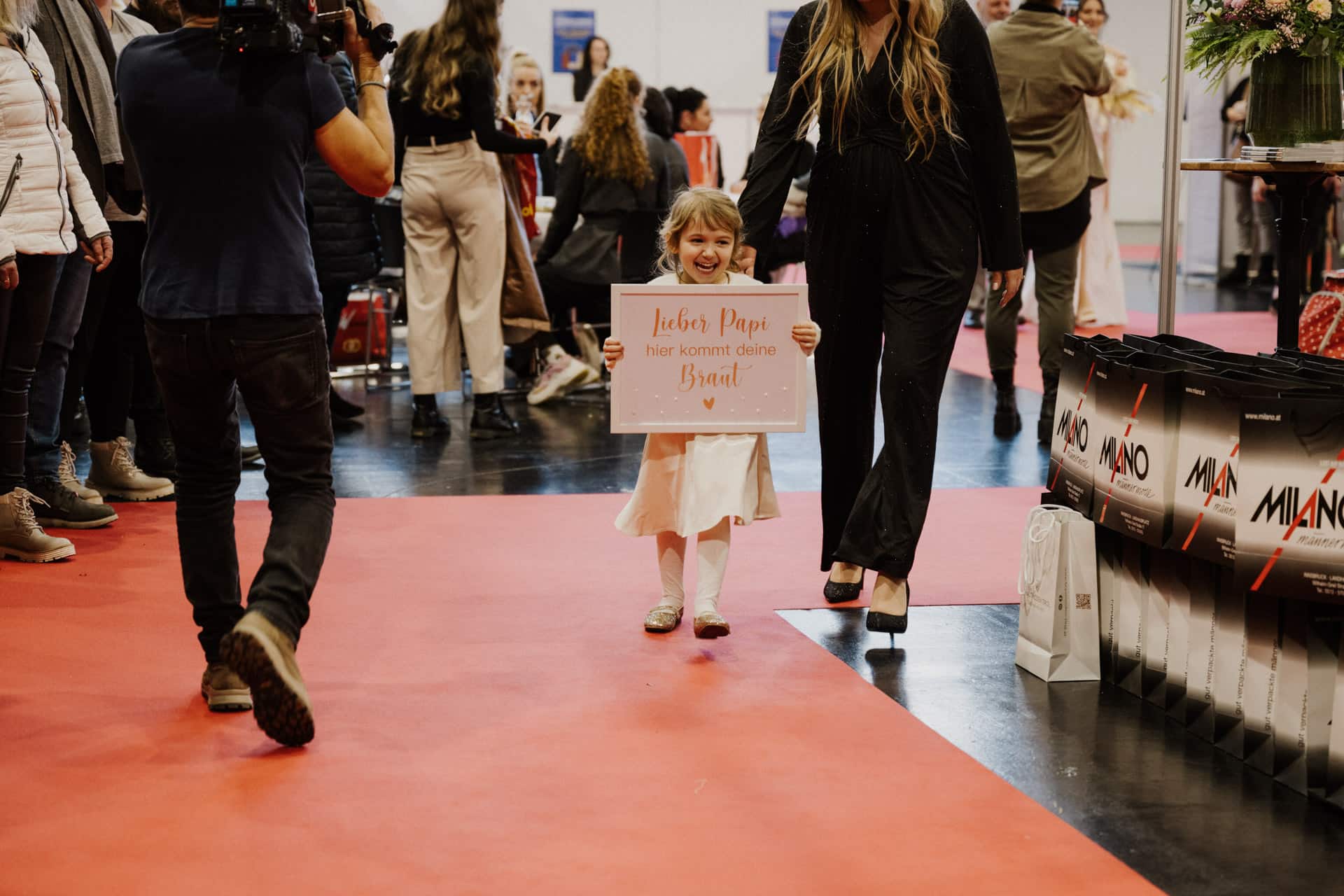 Ein junges Mädchen in einem weißen Kleid läuft bei einer festlichen Veranstaltung über einen roten Teppich und hält ein Schild mit einer Botschaft auf Deutsch in der Hand. Sie lächelt, auf beiden Seiten von Menschen und Fotografen umgeben. Auf einem Tisch in der Nähe stehen Taschen.