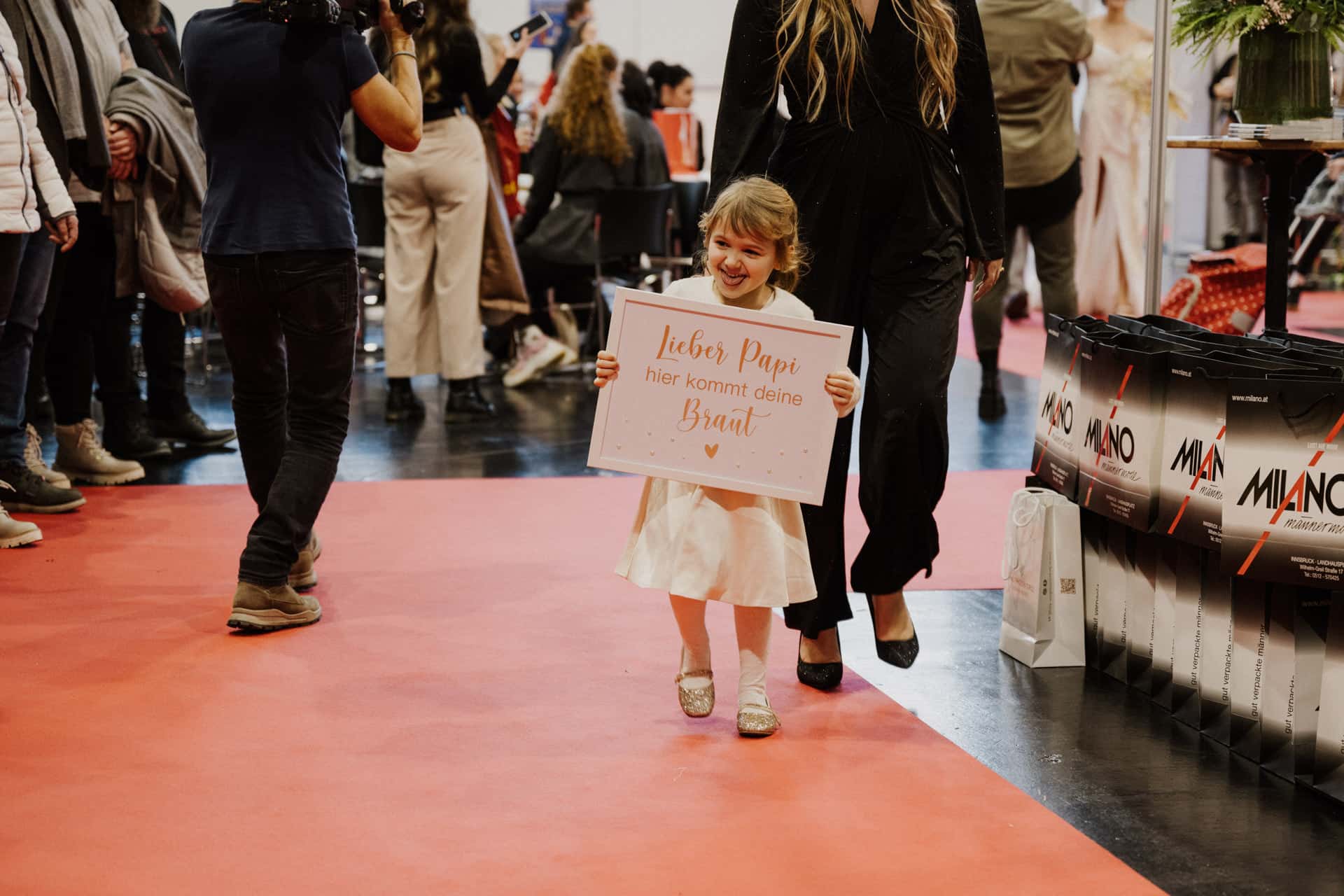 Ein junges Mädchen in einem weißen Kleid schreitet anmutig über einen roten Teppich und hält ein Schild inmitten der eleganten Ausstellungen und der geschäftigen Menge. Die Szene strahlt den festlichen Charme von Hochzeiten & Festen bei einer bezaubernden Veranstaltung oder Ausstellung aus.