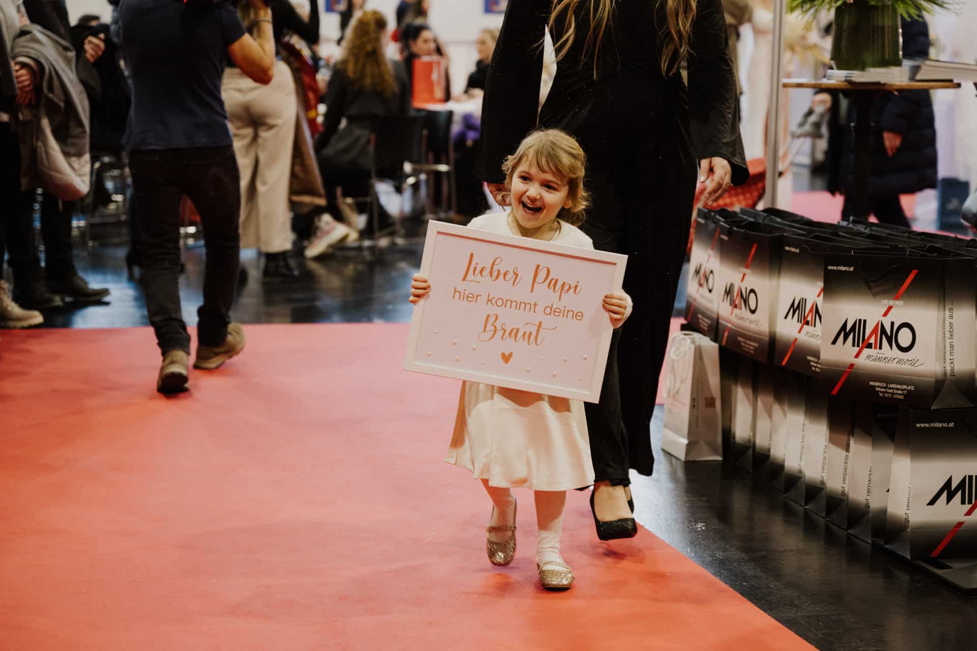 Ein junges Mädchen in einem weißen Kleid läuft über einen roten Teppich und hält ein Schild mit deutscher Aufschrift in der Hand. Im Hintergrund, inmitten der lebhaften Atmosphäre von Hochzeiten und Festen, tummeln sich Menschen um Pappkartons mit der Aufschrift „MILANO“.