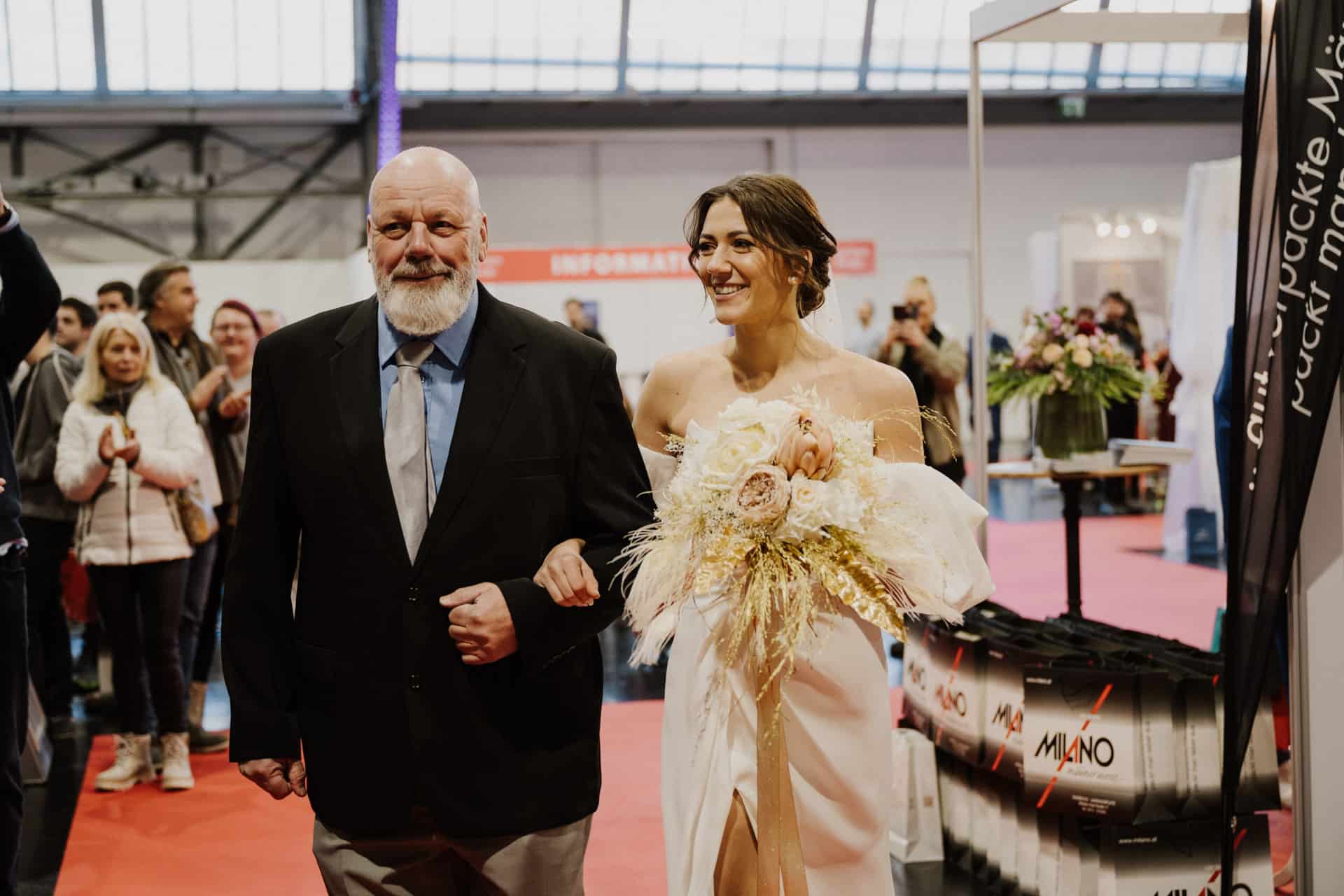 Eine lächelnde Braut in einem weißen Kleid hält einen Blumenstrauß in der Hand und geht Arm in Arm mit einem älteren Mann im Anzug. Sie zieren den roten Teppich bei einer Hochzeit in geschlossenen Räumen, während Zuschauer im Hintergrund den festlichen Moment genießen.