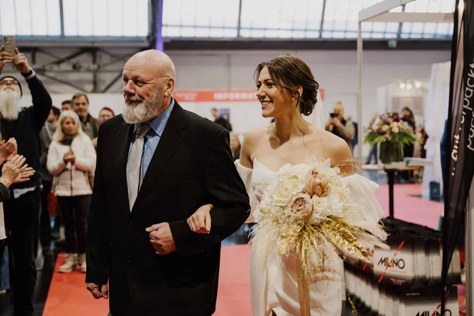 Eine lächelnde Braut in einem weißen Kleid, die die Freude einer Hochzeit verkörpert, hält einen Blumenstrauß in der Hand und geht Arm in Arm mit einem Mann in schwarzem Anzug und blauem Hemd über einen roten Teppich. Zuschauer versammeln sich um sie herum, einige machen Fotos. Der Veranstaltungsort strahlt mit seinen großen Fenstern eine schicke Industrieatmosphäre aus.