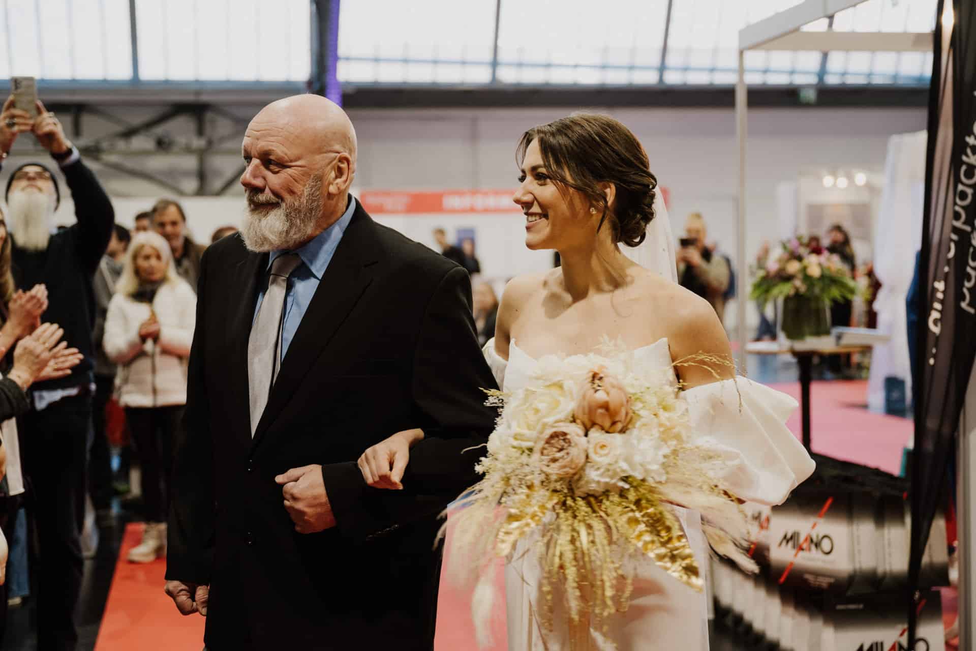 Bei Hochzeiten geht eine Braut in einem weißen Kleid mit einem Blumenstrauß Arm in Arm mit einem kahlköpfigen Mann im schwarzen Anzug. Lächelnd schlendern sie durch den festlichen Innenbereich, im Hintergrund voller Menschen.