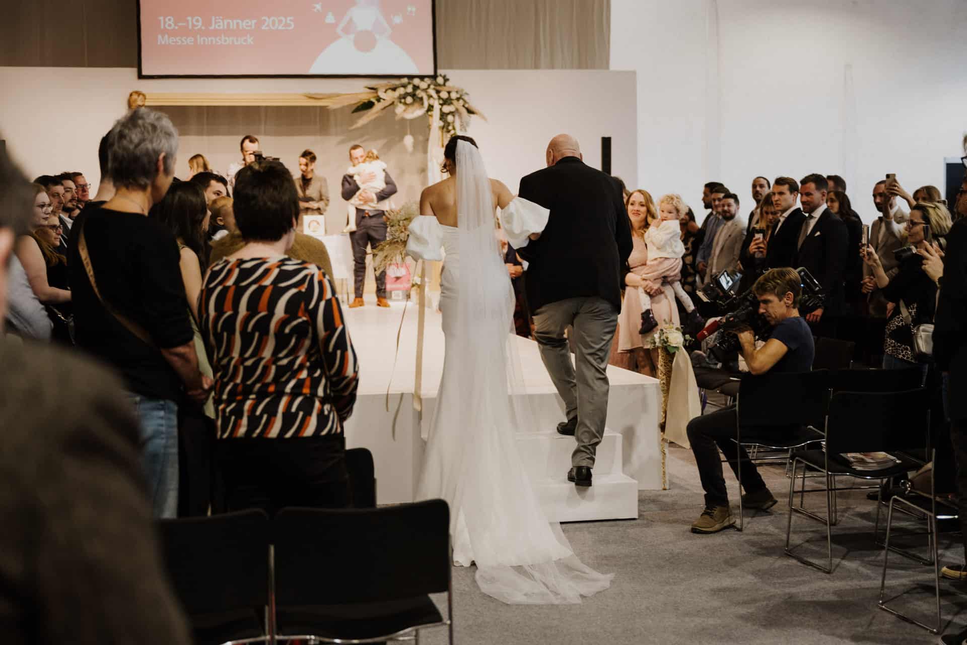 Eine Braut in einem weißen Kleid schreitet bei der Zeremonie mit einem älteren Mann zum Altar und verkörpert damit die Essenz von Hochzeiten & Feste. Die Gäste sitzen oder halten Momente fest, während auf einem Bildschirm Details zu einer Veranstaltung am 18. und 19. Januar 2025 in der Messe Innsbruck angezeigt werden.