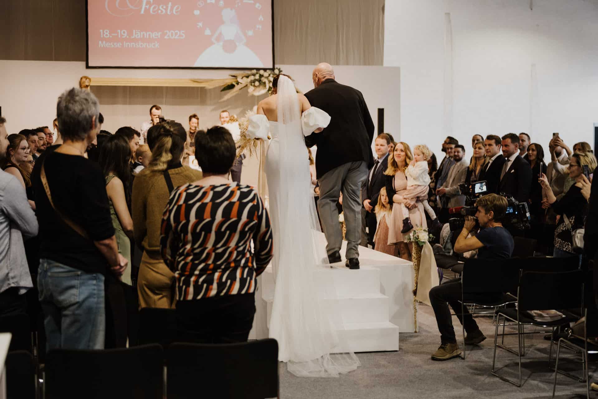 Eine Braut in weißem Kleid und Schleier geht mit einem älteren Mann im dunklen Anzug die Treppe hinauf und blickt auf eine Menschenmenge am Hochzeitsort. Zuschauer sitzen und stehen und fangen die Magie der Hochzeiten ein, während ein Kameramann das Ereignis filmt. Ein Bildschirm im Hintergrund zeigt Daten und Ort an.