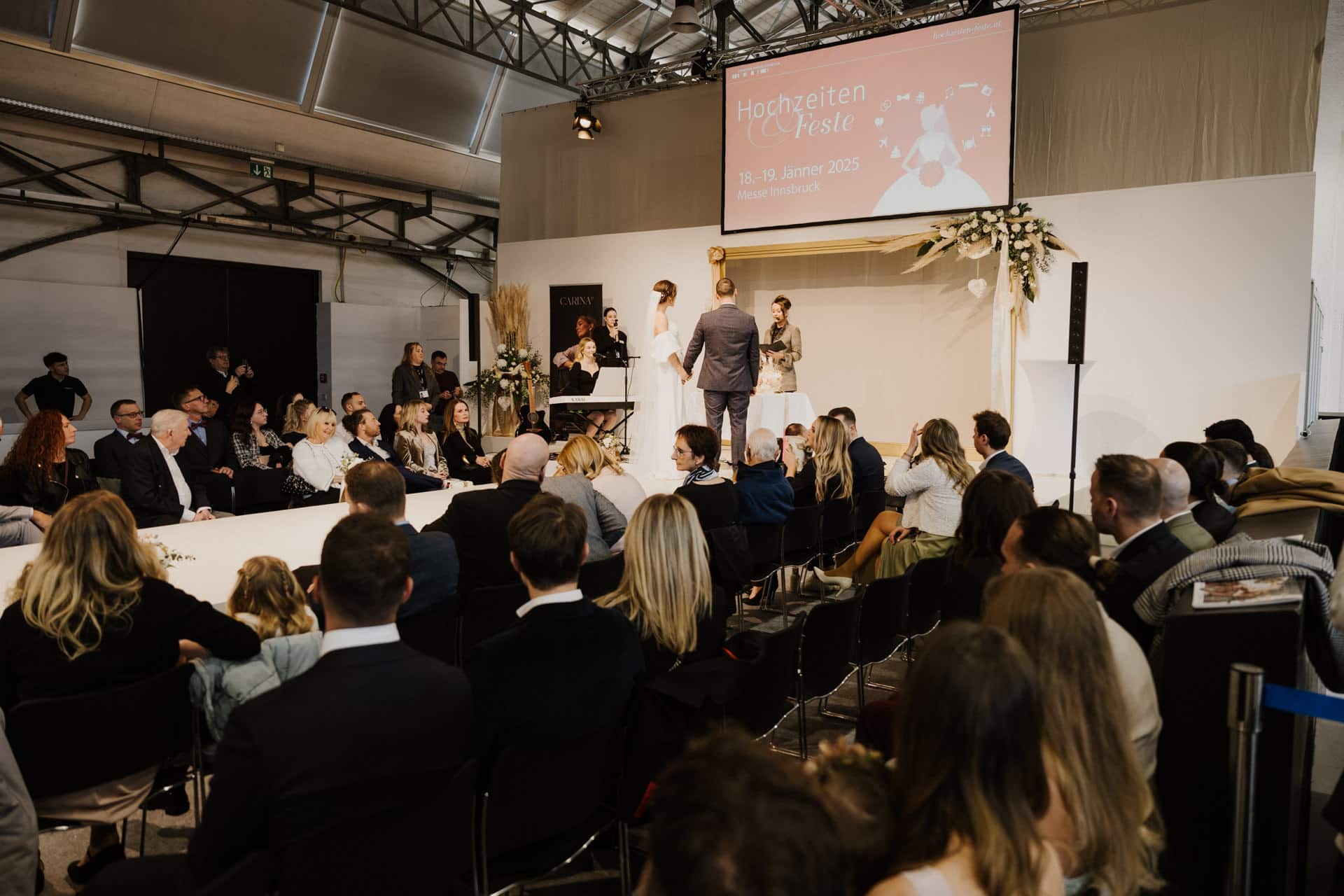 Ein Redner hält auf einer Hochzeitsmesse einen Vortrag. Die Teilnehmer sitzen in Reihen vor der Leinwand, auf der neben den Veranstaltungsdaten prominent „Hochzeiten & Feste“ angezeigt wird. Ein Blumenbogen verleiht dem Vortrag Charme, während die Zuschauer in dem geräumigen, gut beleuchteten Veranstaltungsort aufmerksam zuschauen.