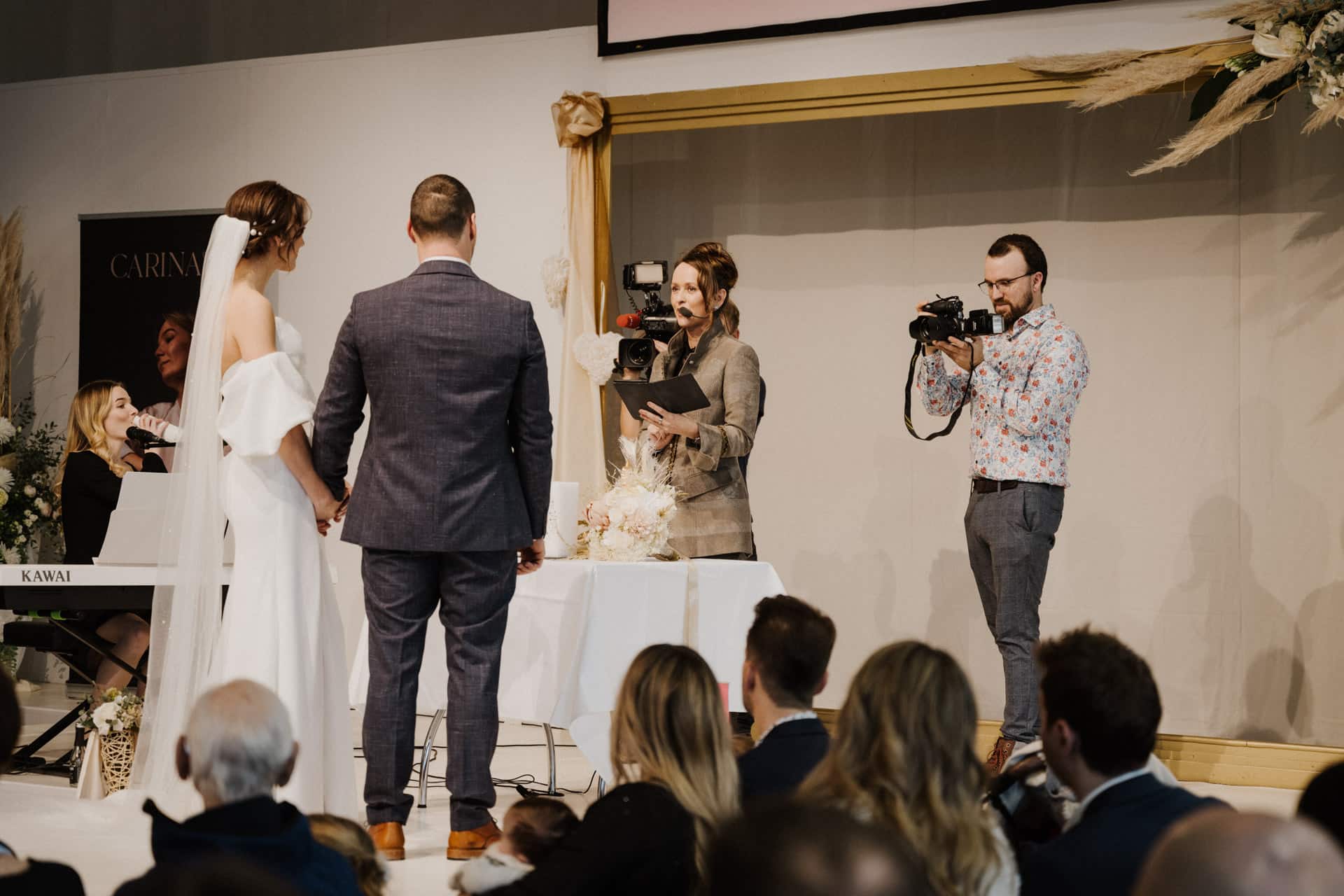 Während einer wunderschönen Hochzeitszeremonie steht das Paar dem Geistlichen gegenüber. Die Braut strahlt in ihrem weißen Kleid und der Bräutigam sieht in seinem grauen Anzug schick aus. Während ein Fotograf diesen besonderen Moment inmitten von Hochzeiten & Feste festhält, spielt ein Musiker Klavier für die sitzenden Gäste.