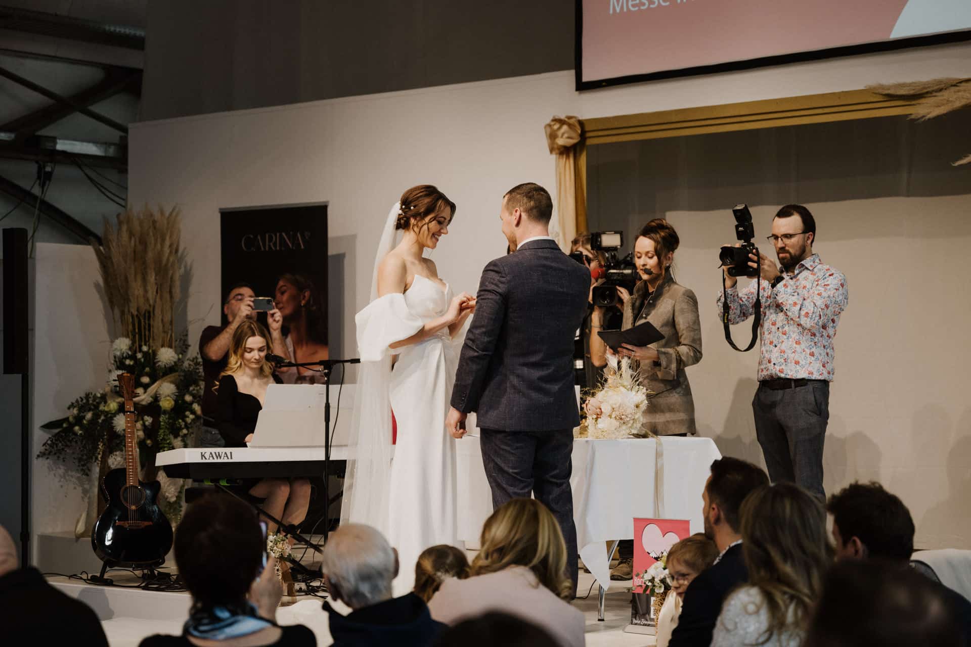 Ein Paar tauscht bei einer Hochzeitszeremonie die Ringe aus, einem Höhepunkt von Hochzeiten & Feste. Ein Pianist spielt in der Nähe, während der Fotograf und die Gäste den Moment einfangen, umgeben von eleganter Dekoration und Blumen.