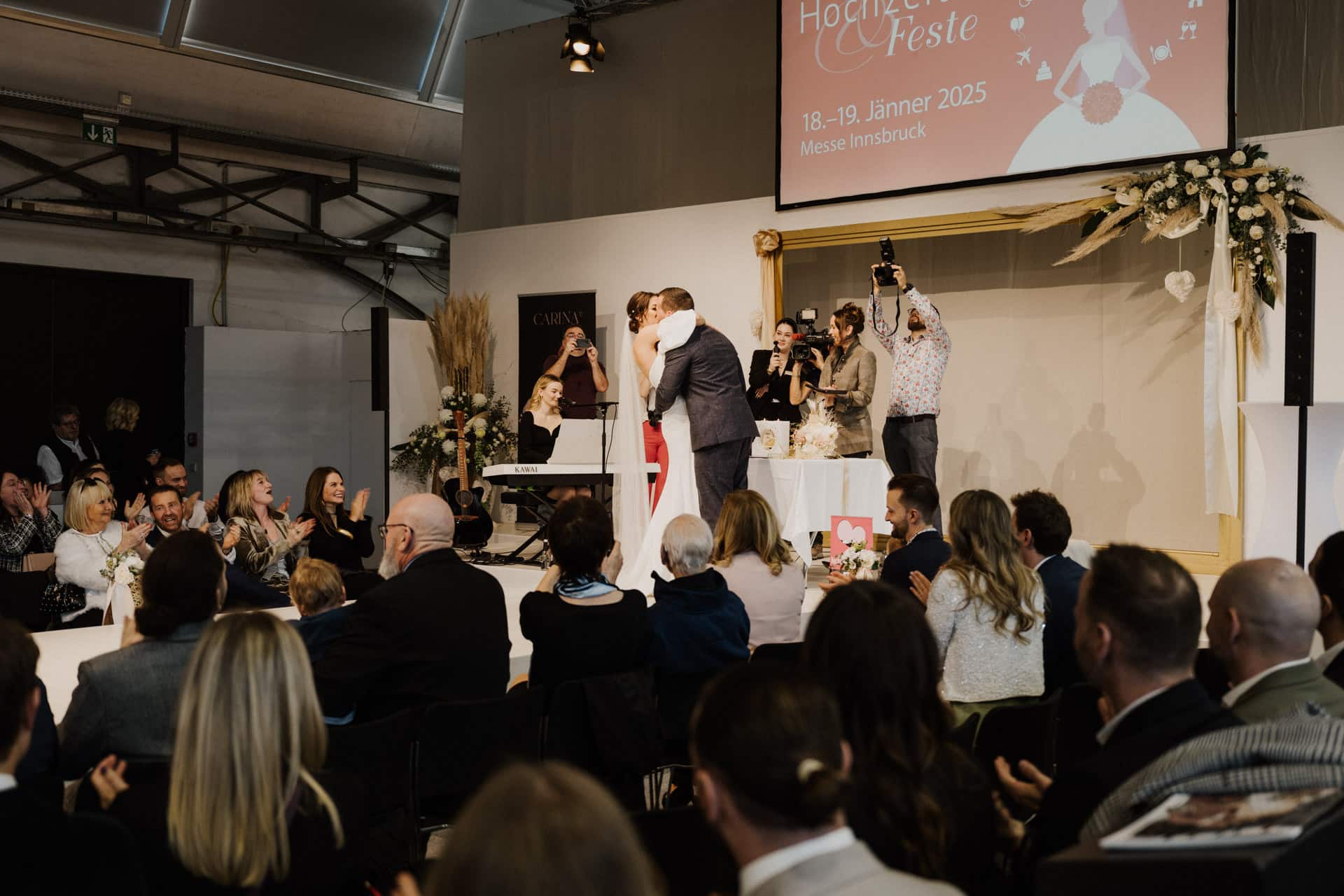 Ein Paar küsst sich auf der Bühne einer Hochzeitsveranstaltung und begeistert das Publikum, das zusieht und applaudiert. Ein Fotograf hält den Moment vor einem Banner mit der Aufschrift „Hochzeiten & Feste, 18.-19. Jänner 2025, Messe Innsbruck“ fest. Blumenschmuck schmückt die Bühne.