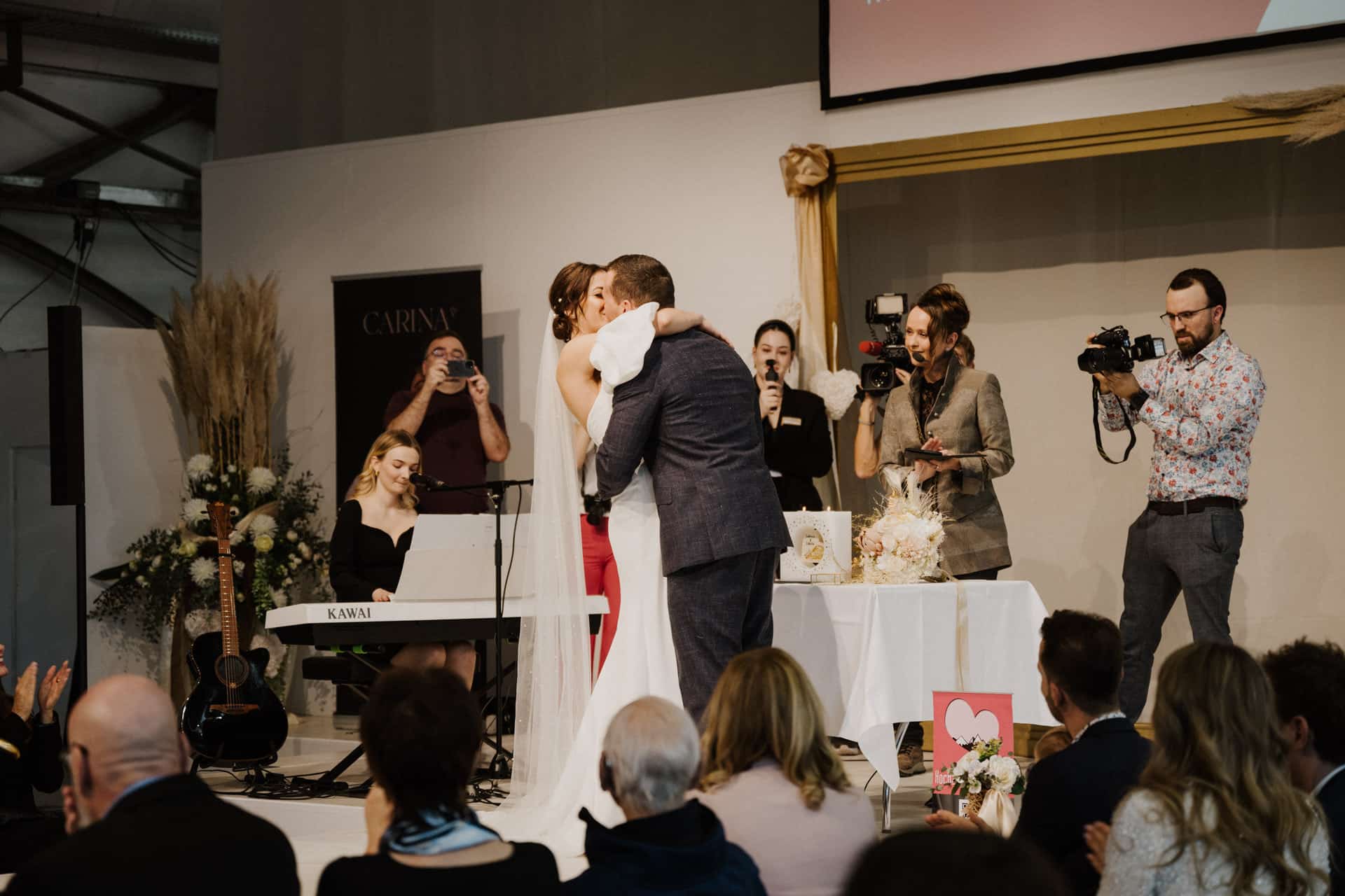 Inmitten der freudigen Feierlichkeiten von Hochzeiten küssen sich Braut und Bräutigam bei ihrer Hochzeitszeremonie zärtlich, umgeben von Gästen. Ein Pianist spielt ein Ständchen, während Fotografen und Zuschauer den Moment festhalten. Blumen und eine dekorative Kulisse rahmen die festliche Szene wunderschön ein.