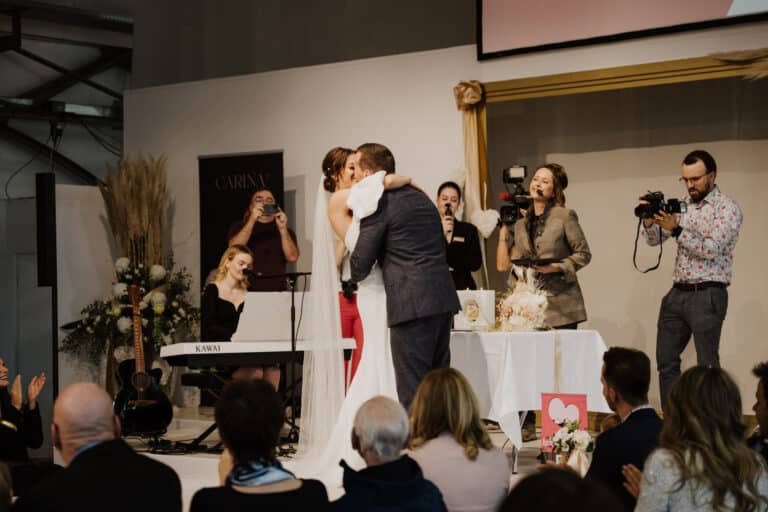 Ein Paar küsst sich während einer Hochzeitszeremonie vor Publikum und verkörpert damit die Freude an Hochzeiten. Eine Frau spielt Keyboard, während Fotografen den Moment festhalten. Ein Blumenarrangement und eine Karte verleihen der Szene auf dem Tisch daneben eine festliche Note.