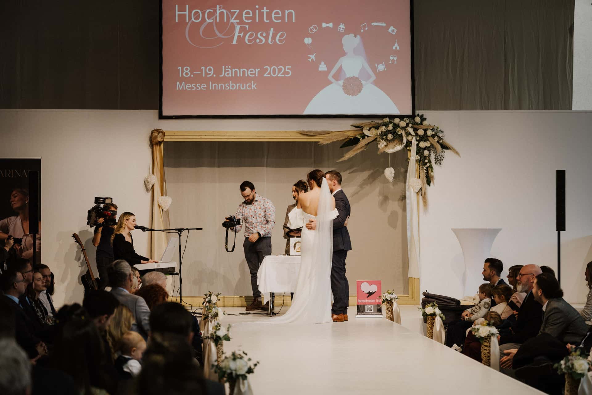 Braut und Bräutigam stehen bei Hochzeiten & Feste am Altar, zu beiden Seiten sitzt das Publikum. Ein Fotograf hält den Moment fest, während die Musiker spielen. Auf einem Bildschirm darüber steht „18.-19. Jänner 2025, Messe Innsbruck“.