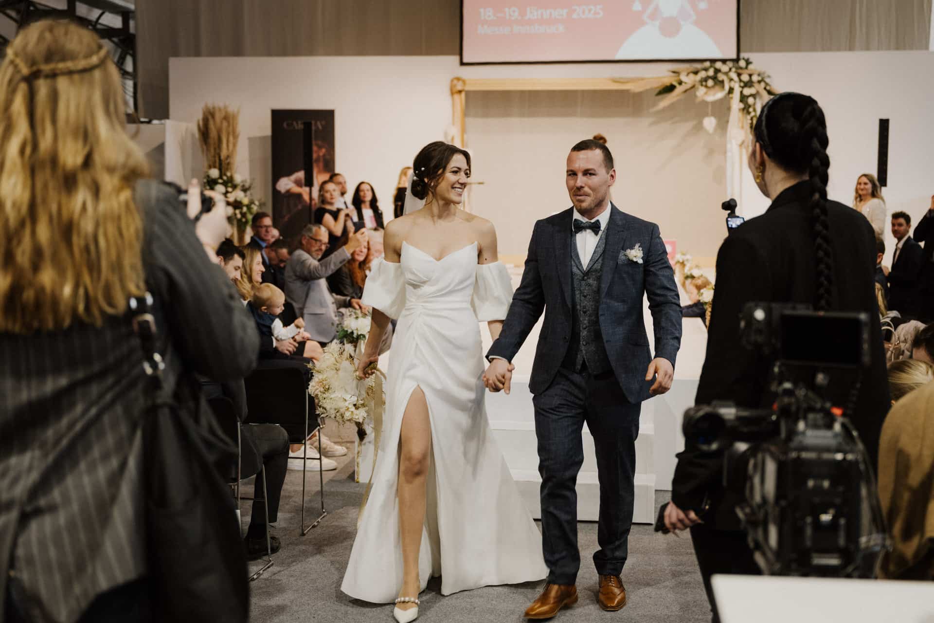 Eine Braut im weißen Kleid und ein Bräutigam im blauen Anzug gehen Hand in Hand den Gang entlang und lächeln herzlich. Gäste sitzen auf beiden Seiten, während der Fotograf diesen freudigen Moment der Hochzeit einfängt. Ein Bildschirm im Hintergrund zeigt Veranstaltungsdetails an und trägt so zur festlichen Atmosphäre bei.