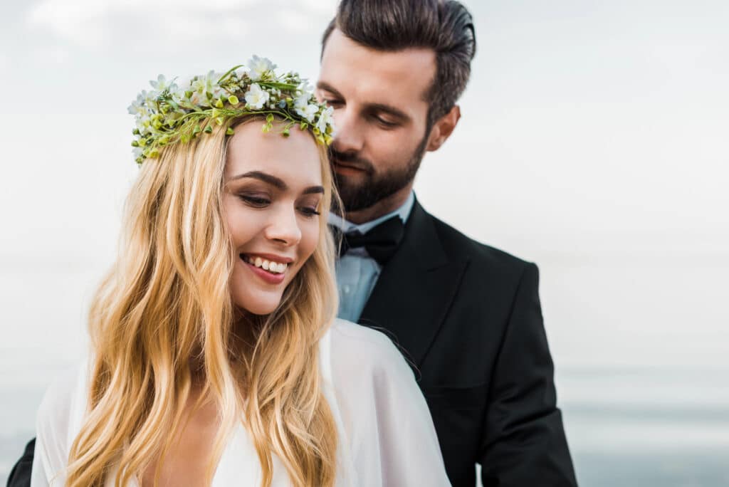 Eine lächelnde Frau mit langen blonden Haaren und einem Blumenkranz steht vor einem Mann im Anzug und verkörpert damit die neuesten Hochzeitstrends. Der Mann mit kurzen dunklen Haaren und Bart ergänzt die Außenkulisse – ein weicher, verschwommener Hintergrund, der Wasser oder Himmel andeutet.