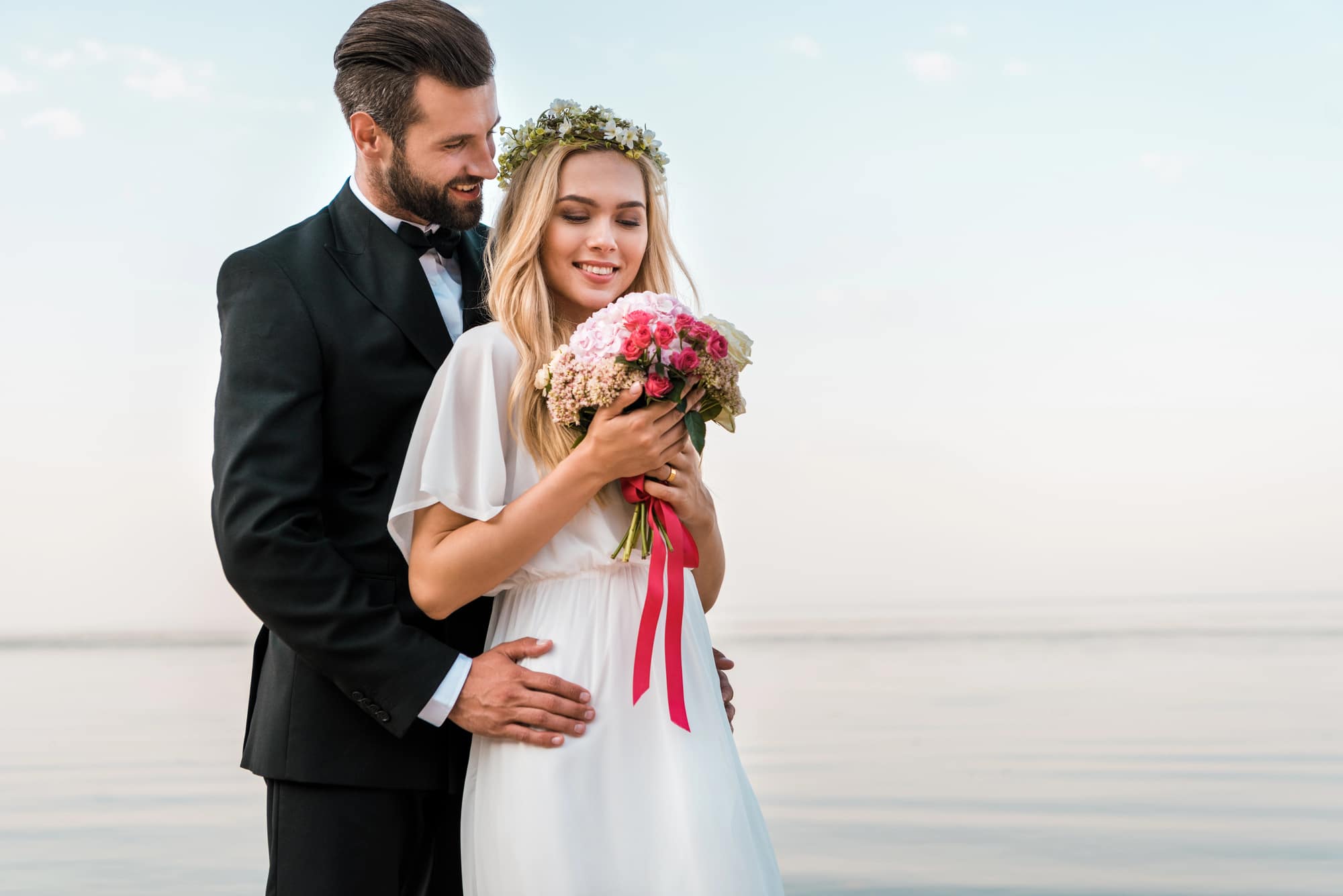 Braut und Bräutigam umarmen sich am Wasser und verkörpern damit die neuesten Hochzeitstrends. Der Bräutigam in einem eleganten schwarzen Anzug hält seine Braut von hinten. Sie strahlt Schönheit aus in ihrem weißen Kleid und Blumenkrone und umklammert einen Strauß rosa Blüten. Sie lächeln sanft unter einem klaren Himmel am ruhigen Wasser.