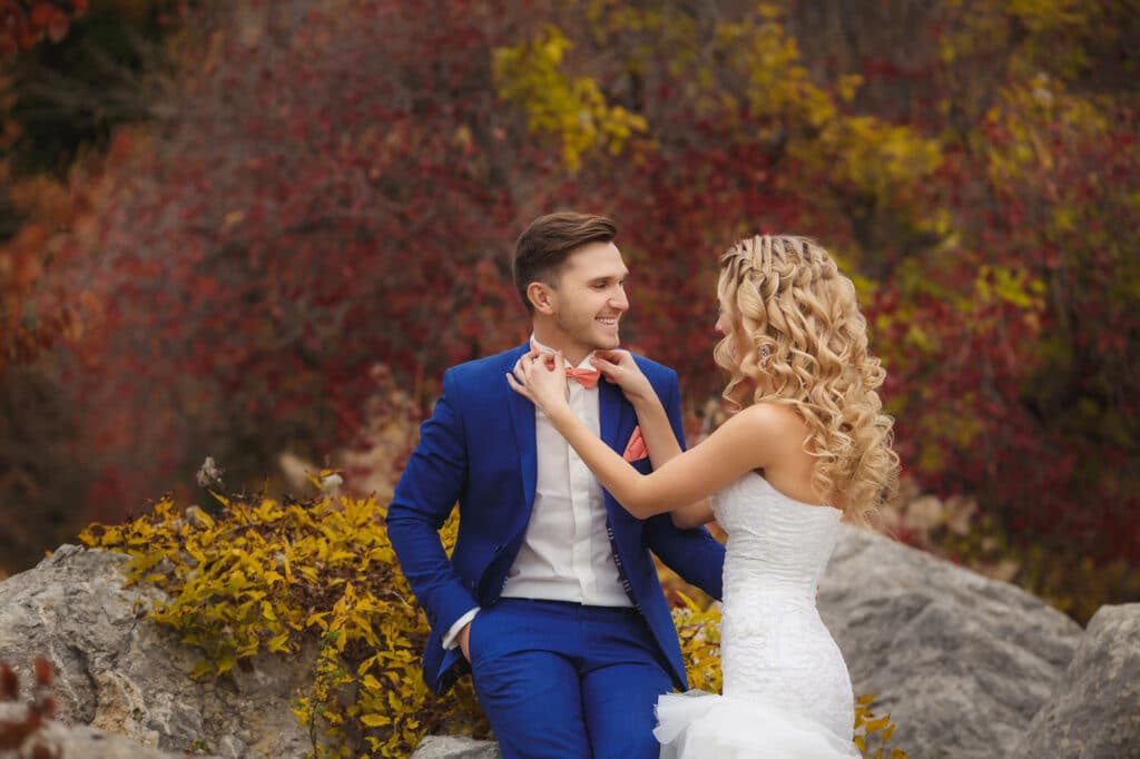 Braut und Bräutigam, die den neuesten Hochzeitstrends folgen, genießen eine malerische Kulisse. Der Bräutigam im blauen Anzug sitzt auf einem Felsen, während die Braut ihre rote Fliege zurechtrückt. Sie sind von herbstlichem Laub in warmen Farben umgeben, das die Essenz moderner Romantik perfekt einfängt.