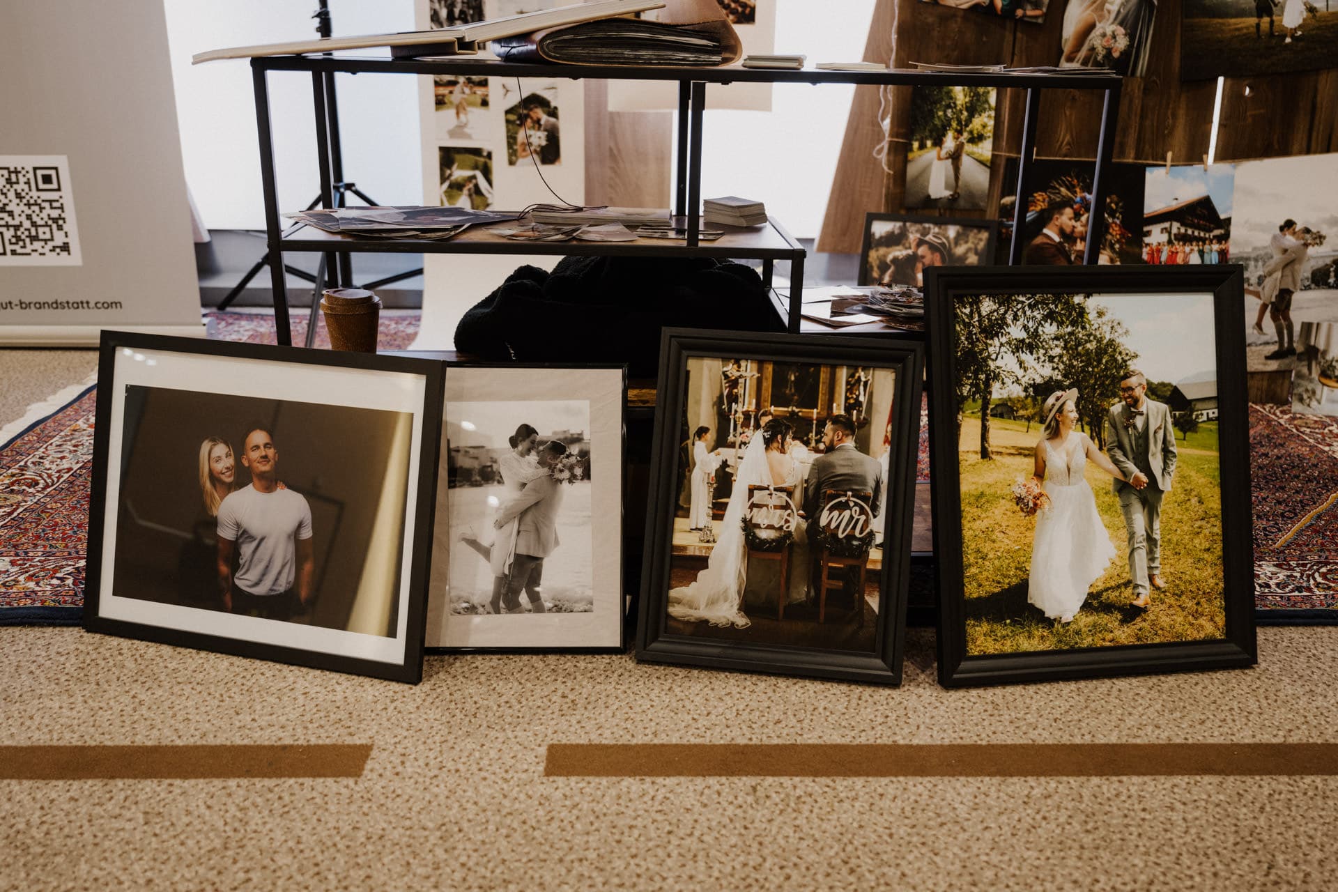 Auf dem Boden stehen gerahmte Fotos, die Lebensfeiern-Momente festhalten: ein Paar, das bei seiner Hochzeit tanzt, Braut und Bräutigam während der Zeremonie und ein Paar, das draußen spazieren geht. Dahinter liegt ein Tisch mit weiteren Fotos und Dekoartikeln.