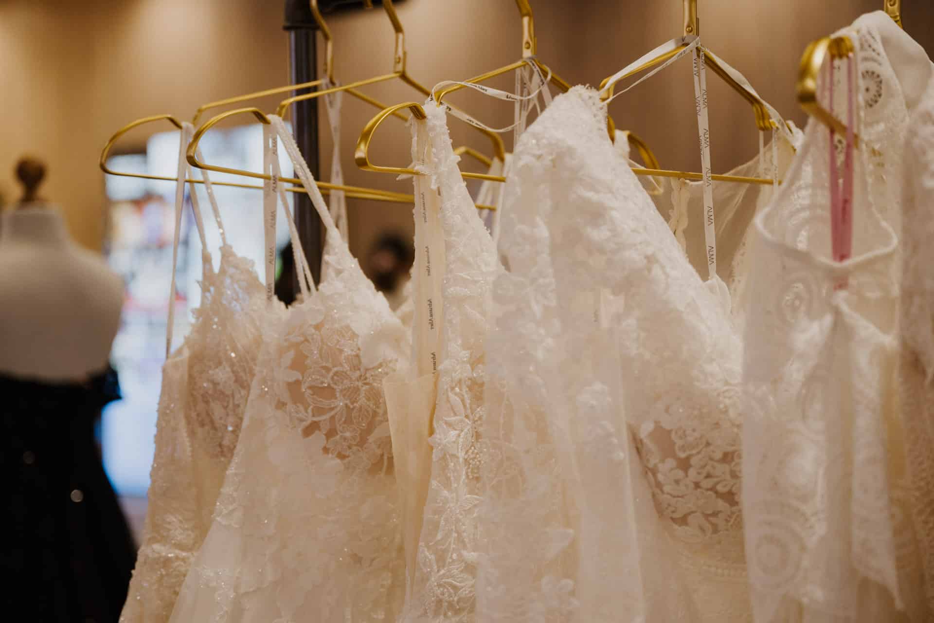 Eine Reihe eleganter Brautkleider aus weißer Spitze auf goldenen Kleiderbügeln, perfekt für Lebensfeiern, ausgestellt in einer Boutique. Der Hintergrund ist sanft verschwommen, wodurch die komplizierten Details und zarten Muster des Stoffes hervorgehoben werden.