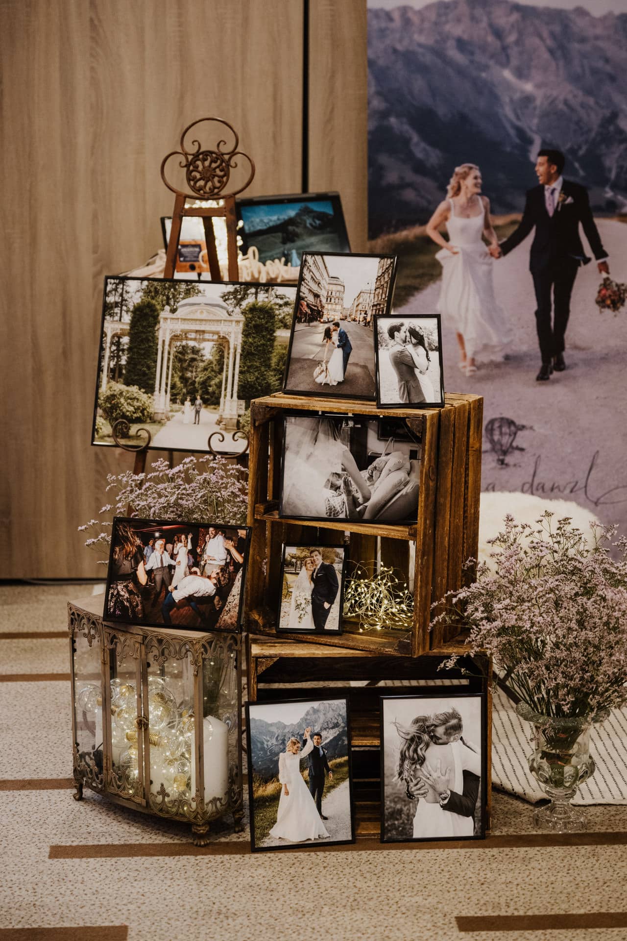 Eine Ausstellung mit Hochzeitsfotos zur Lebensfeier des Paares zeigt gerahmte Bilder, Holzkisten, Blumenarrangements und dekorative Lichter, im Hintergrund ist ein großes Poster zu sehen, auf dem die beiden Hand in Hand gehen.