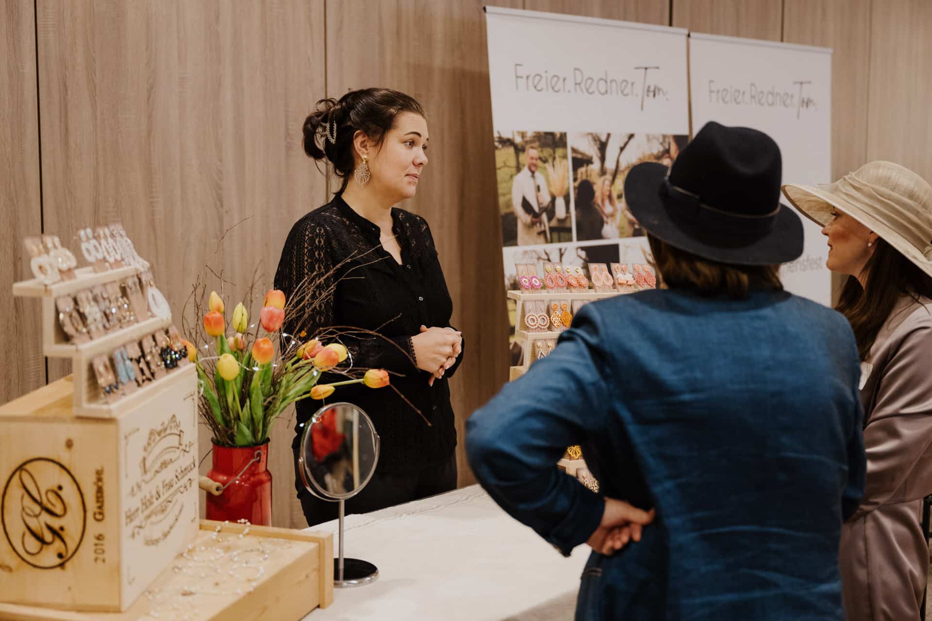 Eine Verkäuferin in einer schwarzen Bluse spricht mit zwei Frauen an einem Marktstand, der Schmuck ausstellt, perfekt für Lebensfeiern. Holzkisten, ein kleiner Spiegel und Tulpen schmücken den Tisch. Die Frauen, die schicke Hüte tragen, begutachten die Stücke, während im Hintergrund Schautafeln mit weiterem Schmuck funkeln.