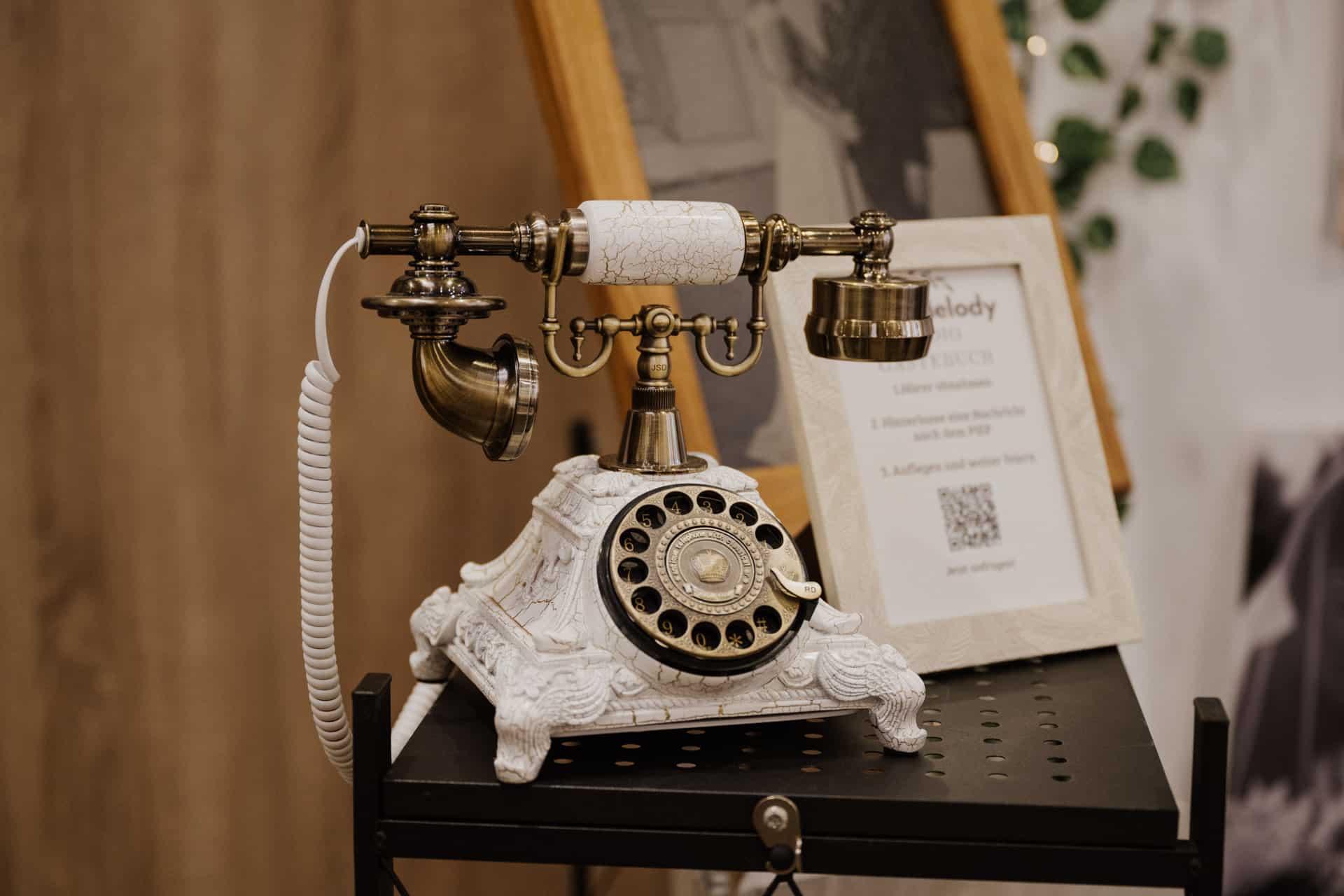 Ein Vintage-Wählscheibentelefon mit verzierten Messing- und Weißdetails steht auf einem schwarzen Metallregal und verleiht der Umgebung einen Hauch von Nostalgie. Im Hintergrund sind dezent ein Informationsschild über Lebensfeiern und ein verschwommenes gerahmtes Foto zu sehen.