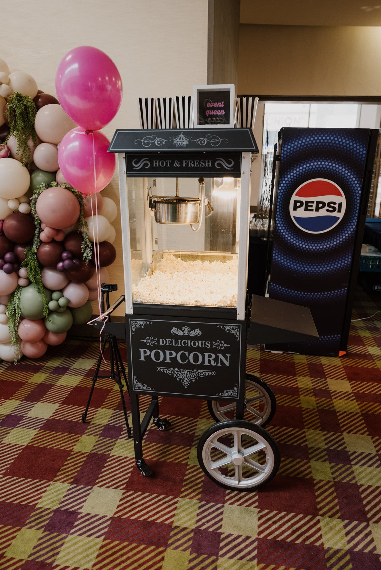 Eine Popcornmaschine mit der Aufschrift „Delicious Popcorn“ steht auf einem gemusterten Teppich neben einem Pepsi-Kühlschrank und feiert Lebensfeiern. Rosa-weiße Luftballons mit einem Blumenarrangement schmücken die linke Seite, während auf einem kleinen Schild an der Maschine „Ginger Queen“ steht.