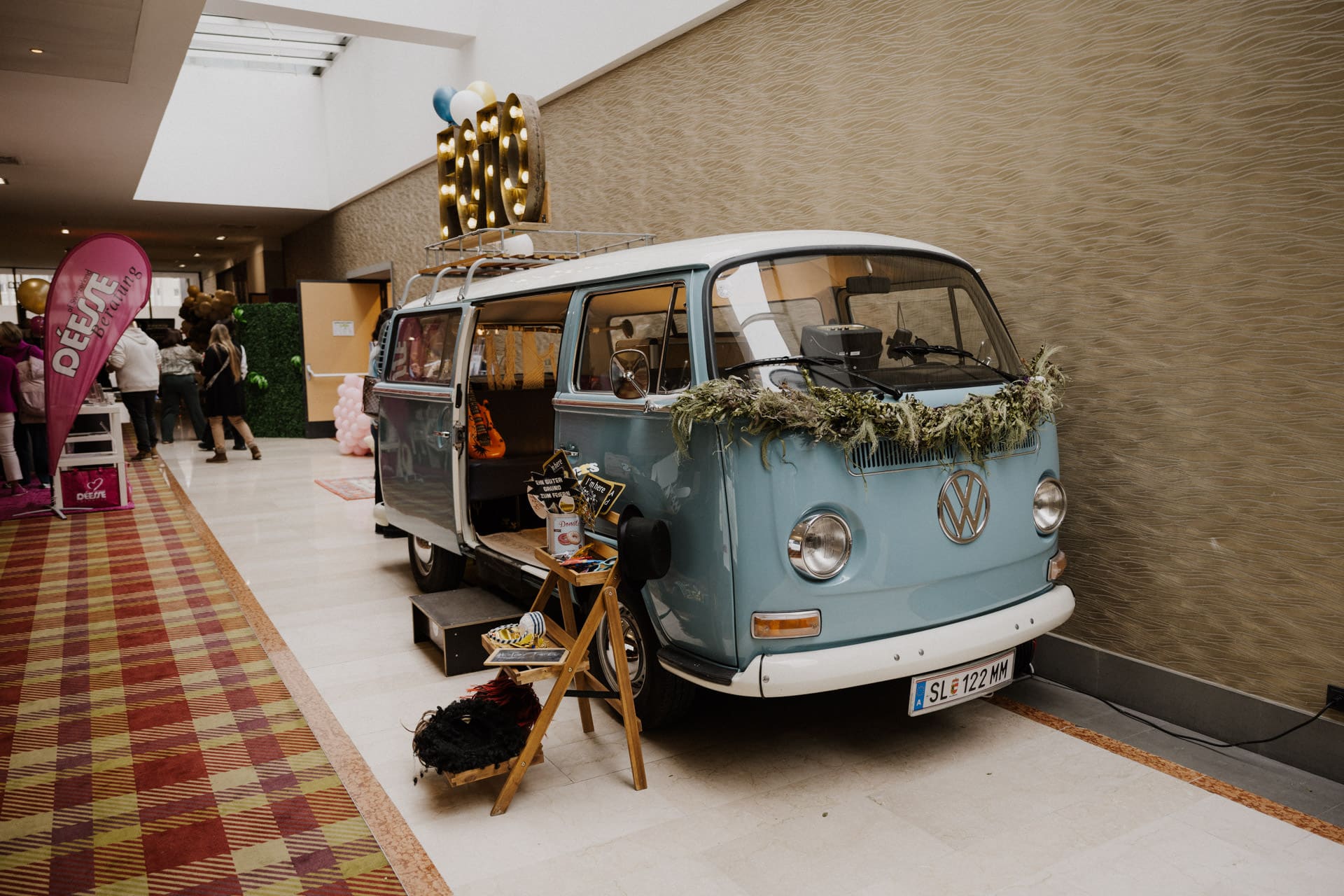 Drinnen parkt ein blau-weißer VW-Bus, geschmückt mit Grünpflanzen und Requisiten, der den Geist der Lebensfeiern einfängt. In der Nähe ist eine kleine Ausstellung mit Fotoautomaten-Artikeln zu sehen, die an eine fröhliche Feier erinnern, die durch festliche Dekorationen noch verstärkt wird.