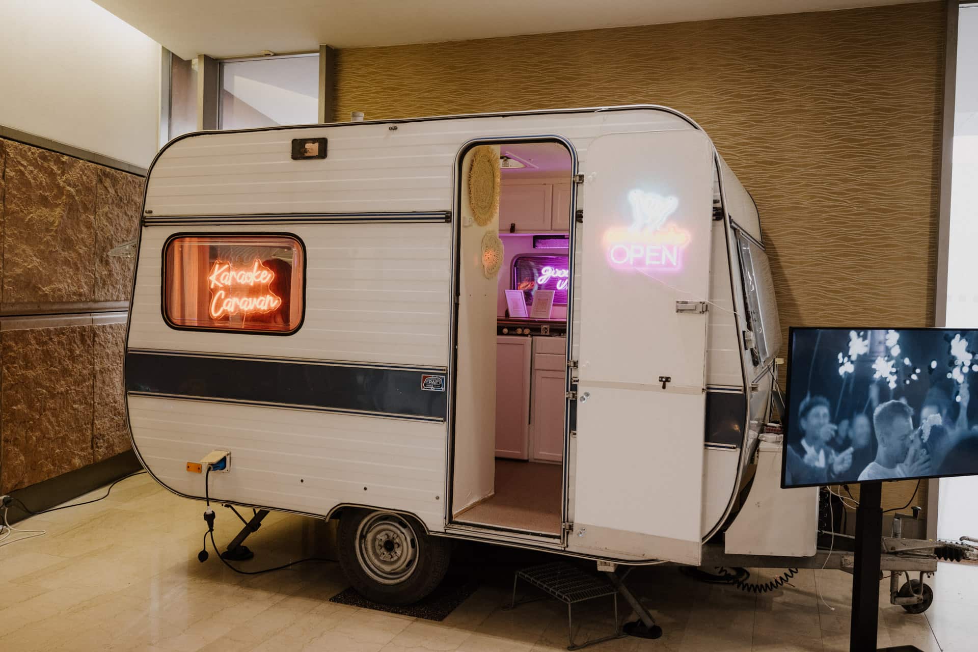 Ein kleiner Oldtimer-Wohnwagen mit einem Neonschild „Karaoke Caravan“ im Fenster und einem „Open“-Schild über der Tür ist bereit für Lebensfeiern. Der Innenraum ist vollgestopft mit Karaoke-Equipment, während draußen eine Leinwand eine lebhafte Menschenmenge zeigt.