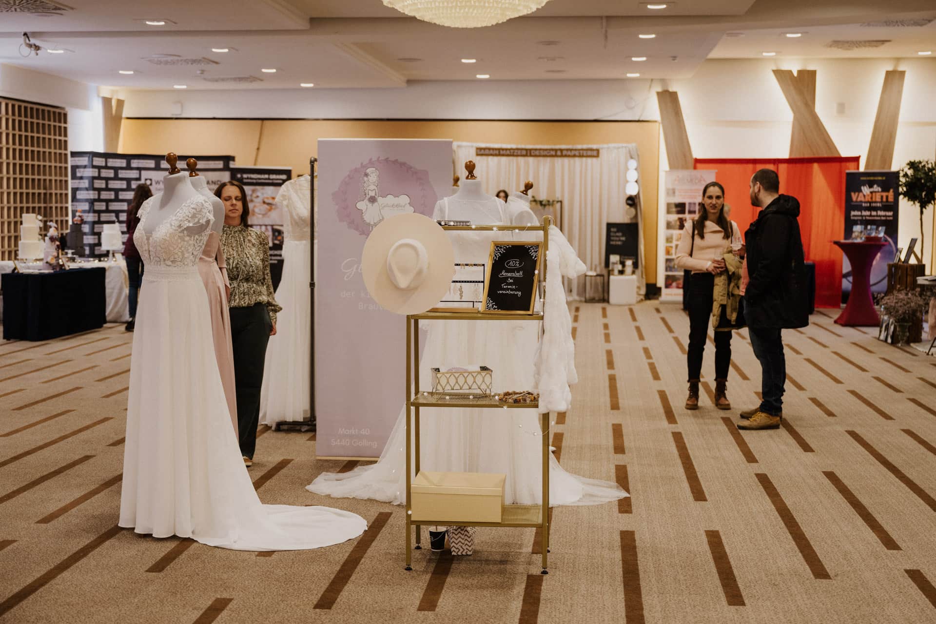 Eine Brautmesse zur Feier der Lebensfeiern präsentiert elegante Brautkleider an Schaufensterpuppen und einen breitkrempigen Hut auf einem Ständer, während die Besucher die Stände durchstöbern. Der geräumige Raum strahlt mit seinen Teppichböden und der geschmackvollen Beleuchtung Charme aus.