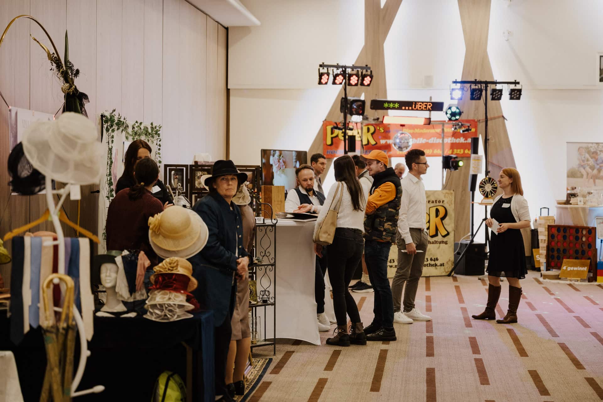 Menschen versammeln sich bei einer Indoor-Veranstaltung mit verschiedenen Ausstellungen, die an eine lebhafte Lebensfeier erinnern. Im Vordergrund sind Hüte und Kleidungsstücke zu sehen, während bunte Banner und Lichter den Hintergrund erhellen. Die Kulisse erinnert an einen lebhaften Marktplatz oder eine Handelsmesse.