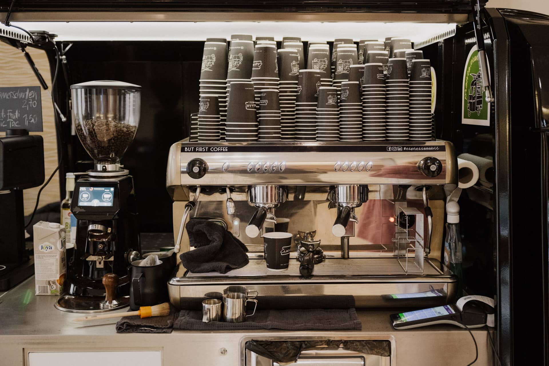 Ein Café-Setup für Lebensfeiern mit einer Espressomaschine, auf der sich ein Stapel schwarzer und weißer Pappbecher befindet. Links davon steht eine Kaffeemühle voller Bohnen. Verschiedene Werkzeuge, Tassen und ein Smartphone sind ordentlich auf der Theke darunter angeordnet und bereit, die feierlichen Momente des Lebens zu servieren.