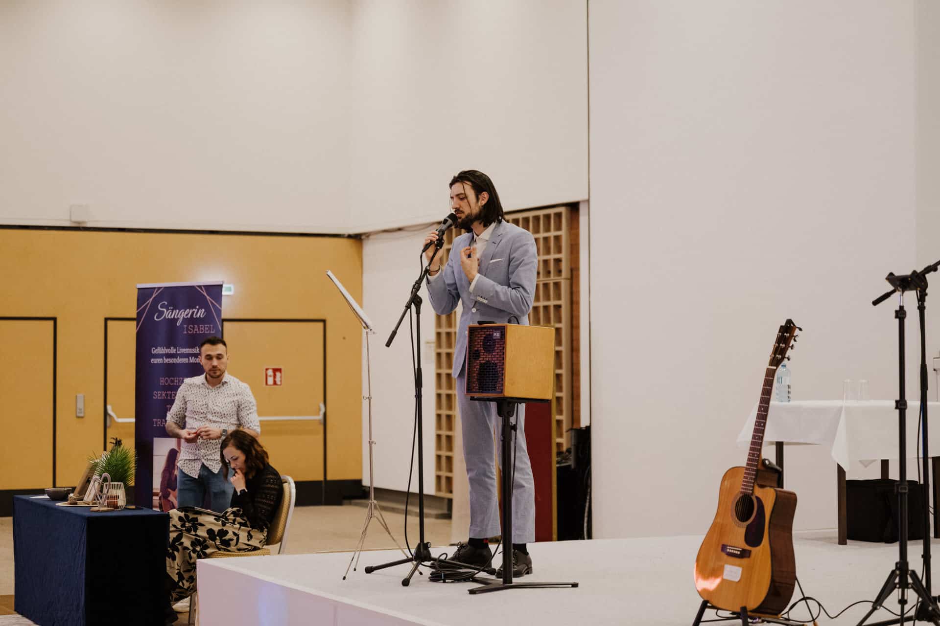 Ein Mann in einem hellblauen Anzug spricht während der Lebensfeiern-Veranstaltung auf der Bühne in ein Mikrofon. Neben ihm steht eine Gitarre, während zwei Personen an einem Tisch mit einer dunklen Tischdecke miteinander interagieren, einer sitzt und einer steht. Im Hintergrund sind ein Banner und einige Klappstühle zu sehen.