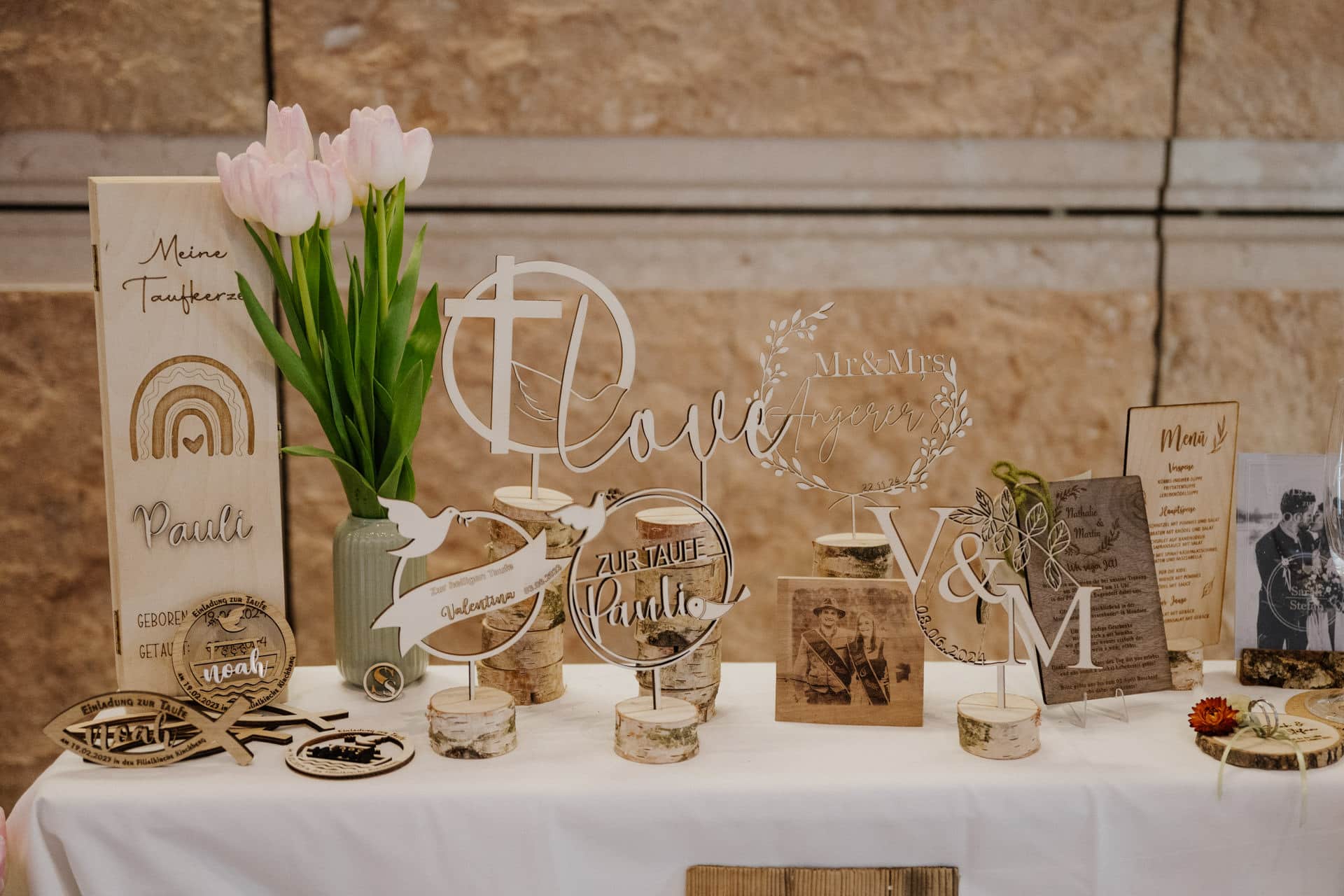 Ein dekorativer Tischaufsteller mit Holz- und Acrylschildern, einer Vase mit rosa Tulpen und verschiedenen Ornamenten feiert Lebensfeiern. Die Gegenstände enthalten eingravierte Botschaften und Symbole zum Thema Liebe und Freude, alles vor einer Steinmauer im Hintergrund.