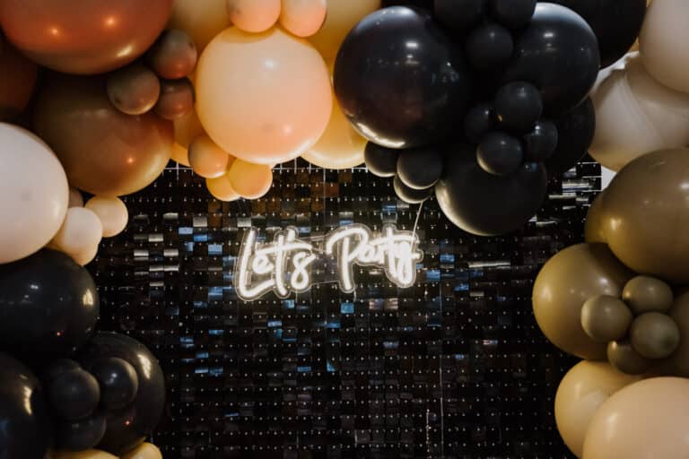 Ein Neonschild mit der Aufschrift „Let’s Party“ ist von einem Bogen aus großen Luftballons in den Farben Schwarz, Gold und Pfirsich umgeben. Der Hintergrund, der den Geist von Lebensfeiern verkörpert, besteht aus einer Wand mit einem schimmernden Design.