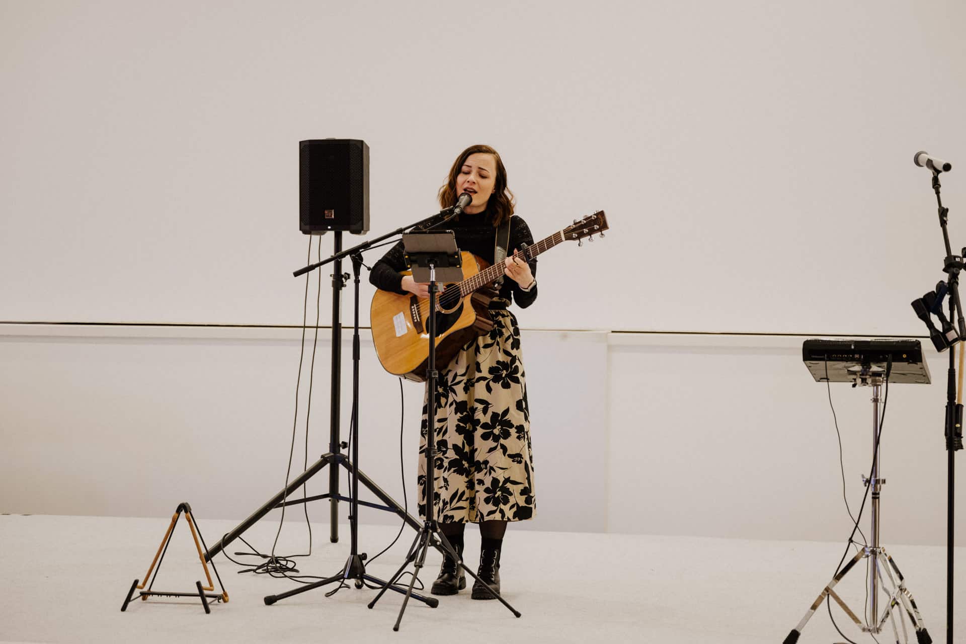 Eine Frau tritt auf der Bühne auf, singt und spielt Akustikgitarre. Sie steht vor einem Mikrofonständer, daneben ein Lautsprecher und ein Notenständer, was die intime Atmosphäre einer Lebensfeier vermittelt. Der Hintergrund ist schlicht weiß. Sie trägt einen geblümten Rock und wirkt konzentriert.