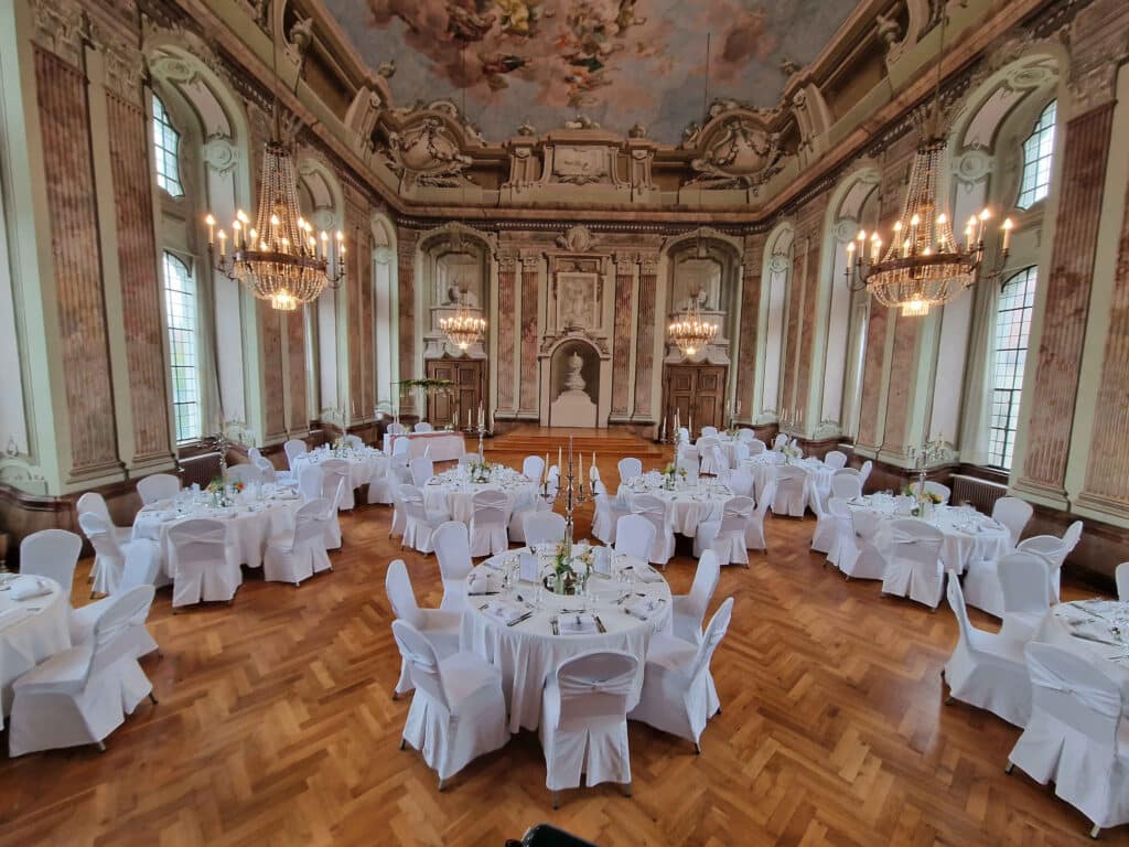 Ein großer und kunstvoller Ballsaal für eine Hochzeitsveranstaltung mit mehreren runden Tischen mit weißen Tischdecken und Stühlen. Erhabene Kronleuchter hängen von einer hohen, verzierten Decke. Hohe Fenster und aufwendige Wanddetails tragen zur eleganten Atmosphäre bei, perfekt für die Hochzeitsplanung.