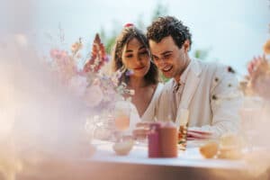 Ein Paar sitzt eng beieinander an einem wunderschön dekorierten Tisch mit Blumen und Getränken und strahlt Freude aus, während sie ein sonniges Ereignis im Freien genießen. Ihre Lächeln lassen darauf schließen, dass es sich um eine Feier handeln könnte, möglicherweise im Zusammenhang mit Hochzeitsfeierlichkeiten oder den Anfängen einer freudigen Hochzeitsplanung.