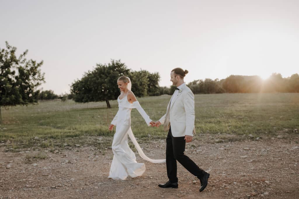 Ein Paar in eleganter Hochzeitskleidung geht Händchen haltend auf einem Kiesweg. Die Braut trägt ein fließendes weißes Kleid und der Bräutigam einen hellen Anzug. Sie sind von einer Wiese mit Bäumen umgeben und im Hintergrund geht die Sonne unter.