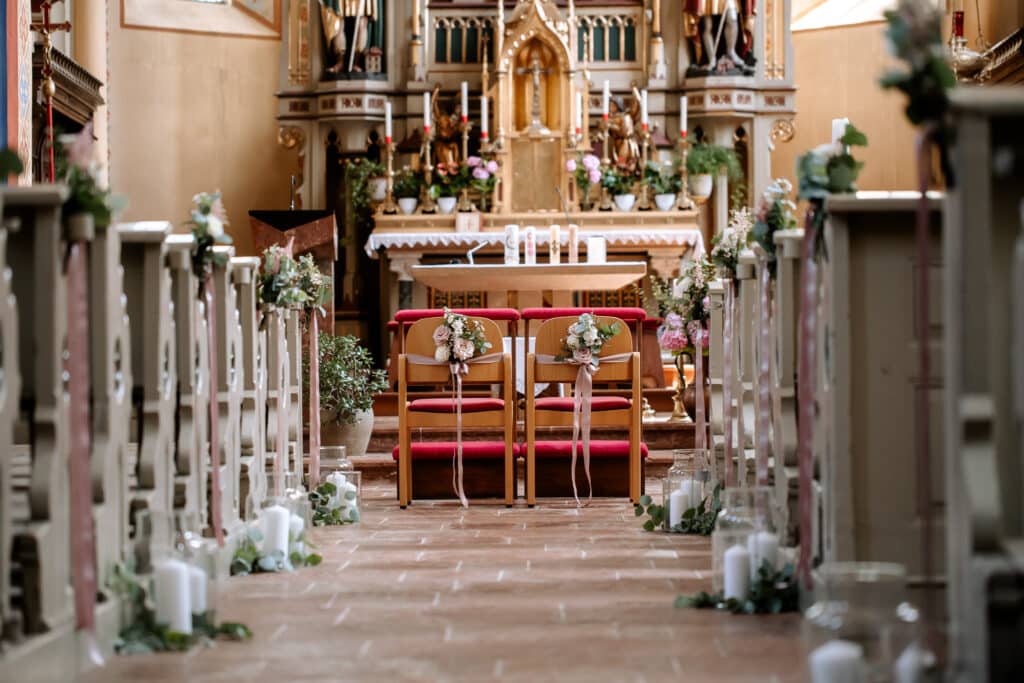 Ein mit weißen Kerzen und Grün geschmückter Kirchengang führt zum Altar. Zwei dekorierte Stühle stehen vor dem Altar, der wunderschön mit Blumen, Kerzen und religiösen Symbolen dekoriert ist. Sonnenlicht fällt durch die Fenster und schafft eine heitere Atmosphäre.