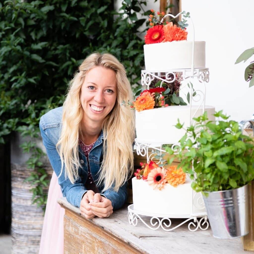 Eine lächelnde Frau mit langen blonden Haaren lehnt an einem Holztisch neben einer mehrstöckigen Torte, die mit roten und orangefarbenen Blumen dekoriert ist. Sie trägt eine Jeansjacke und in der Nähe stehen grüne Pflanzen.